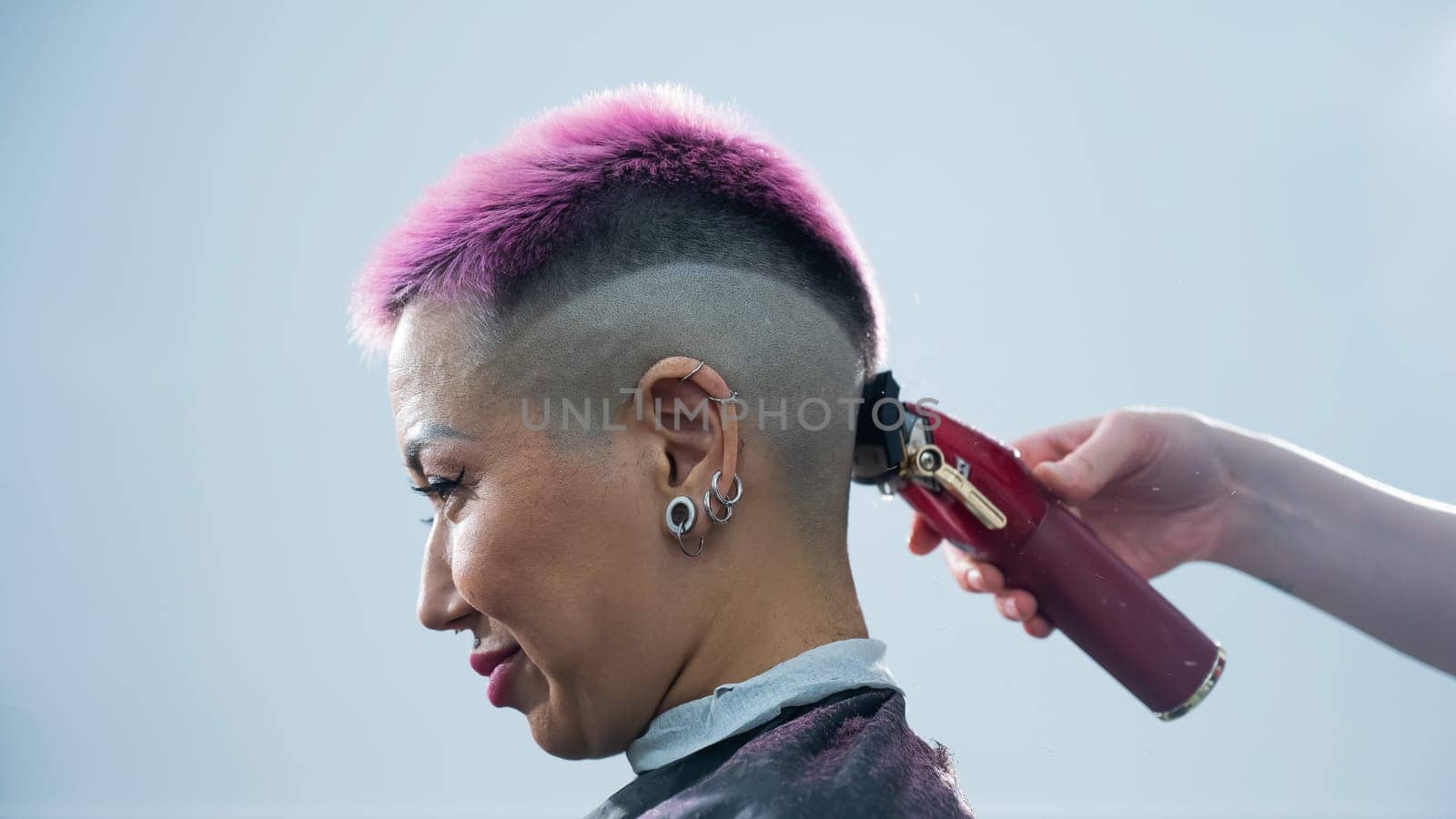 The hairdresser shaves the back of the head of a female client. Rear view of a woman with short pink hair in a barbershop