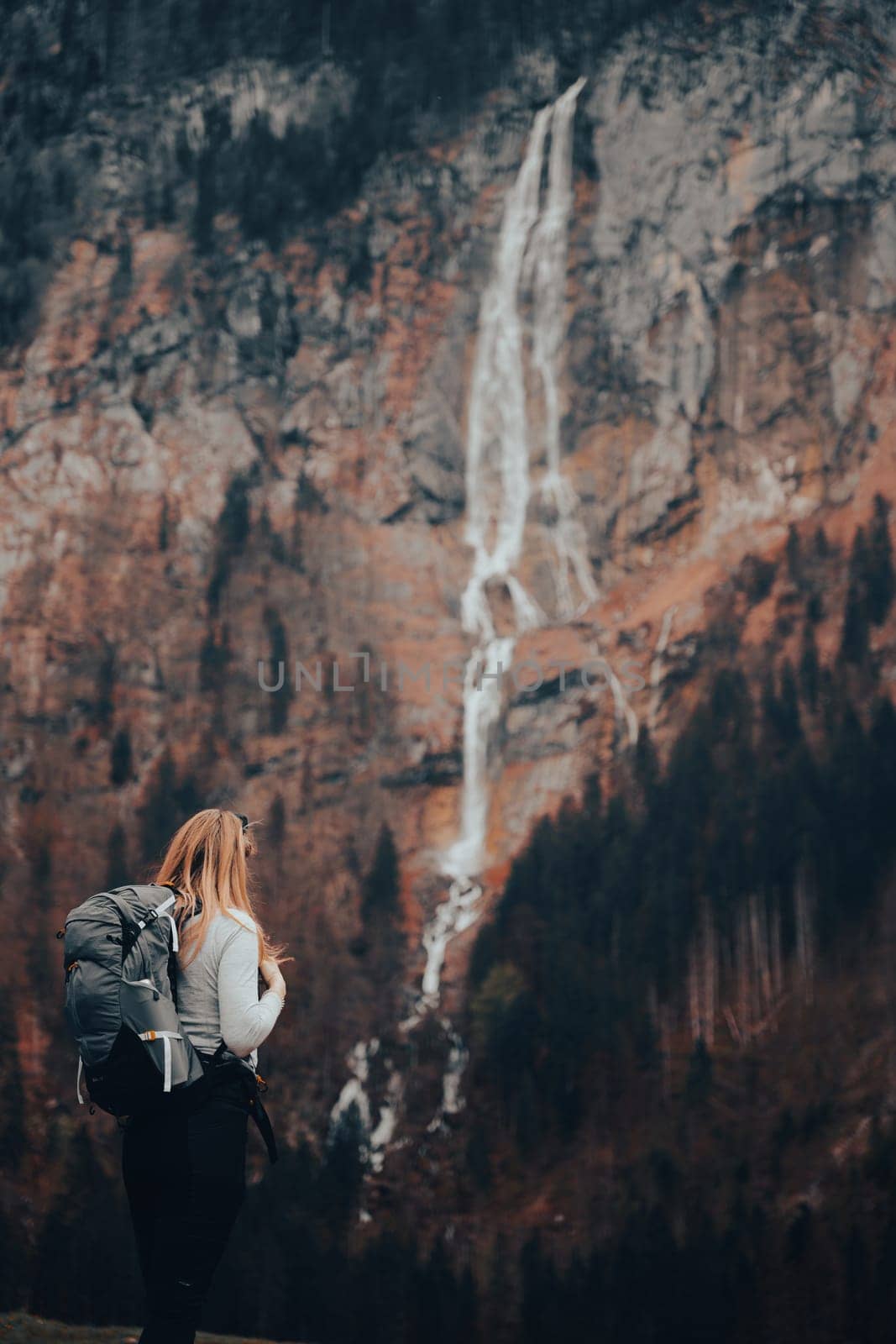 A blond-haired girl is exploring the Austrian Alps. She is in trekking clothes, and with a backpack. She stands with her back to the camera. Looks at the waterfall and holds her hands on the harnesses of her backpack.