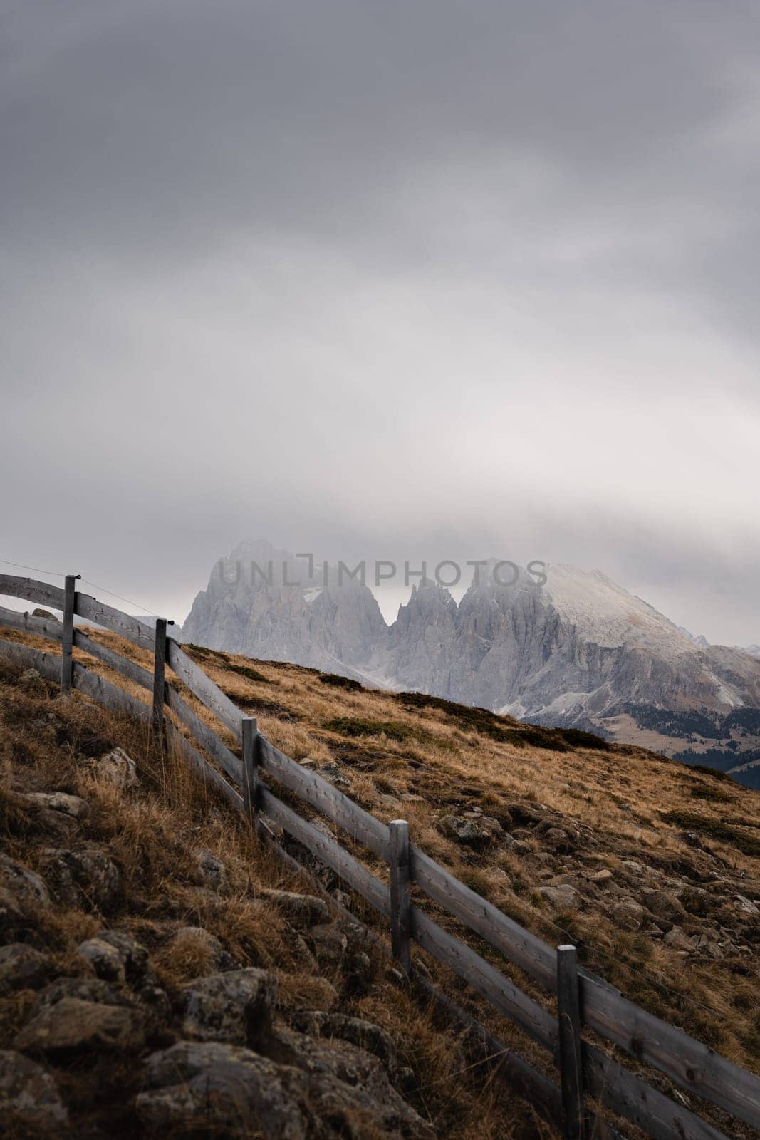 Vertical shot of the autumn moody cloudy alpine landscape with the mountains seen over the hill in the background and a fence for village animals in the foreground. View of the Rosengarten group. Shot made on the hike in Alpe di Siusi, Dolomites, Italy. High-quality photo.