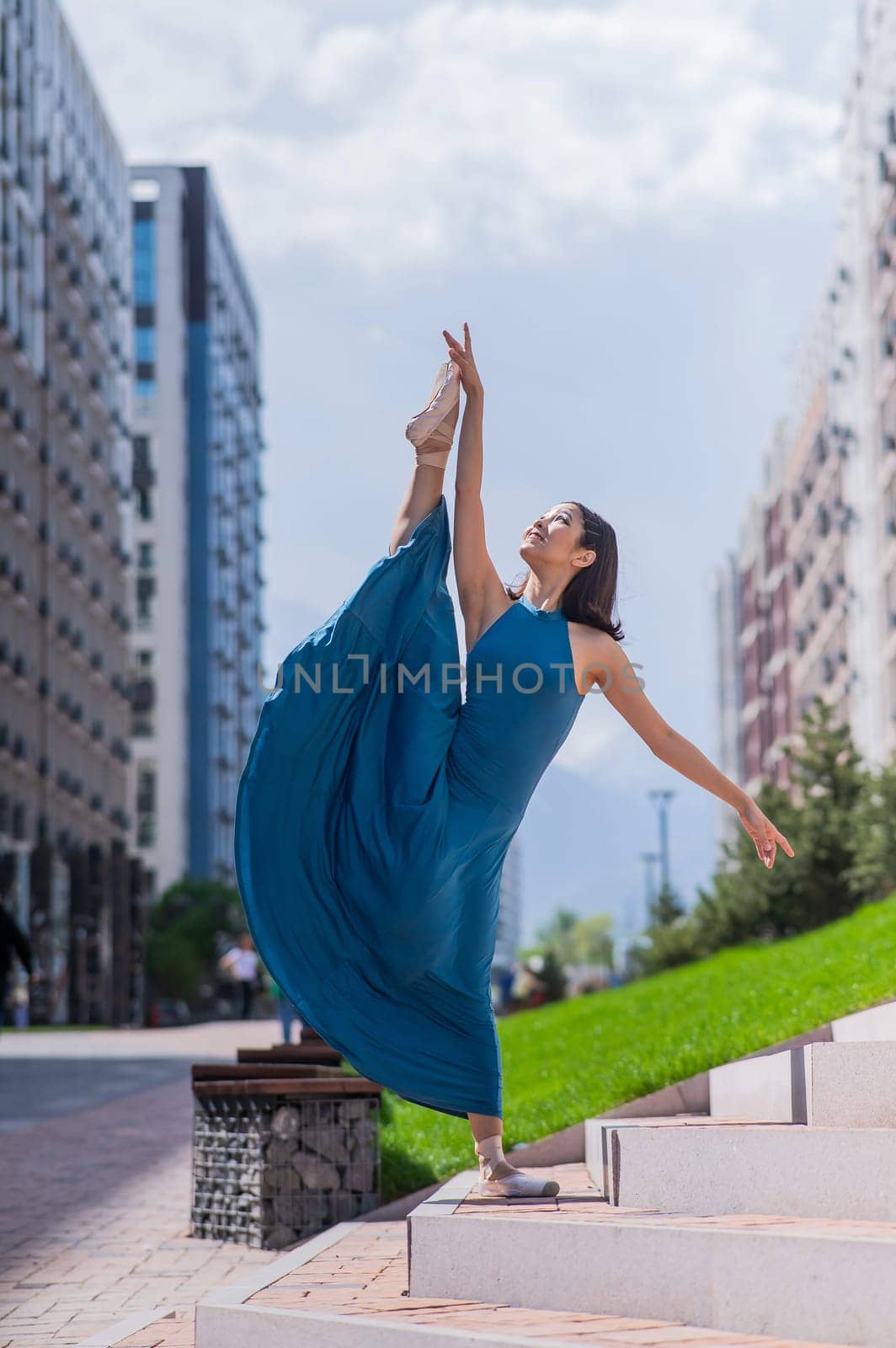 Beautiful Asian ballerina in a blue dress stands on the stairs in the splits outdoors. Urban landscape. Vertical photo