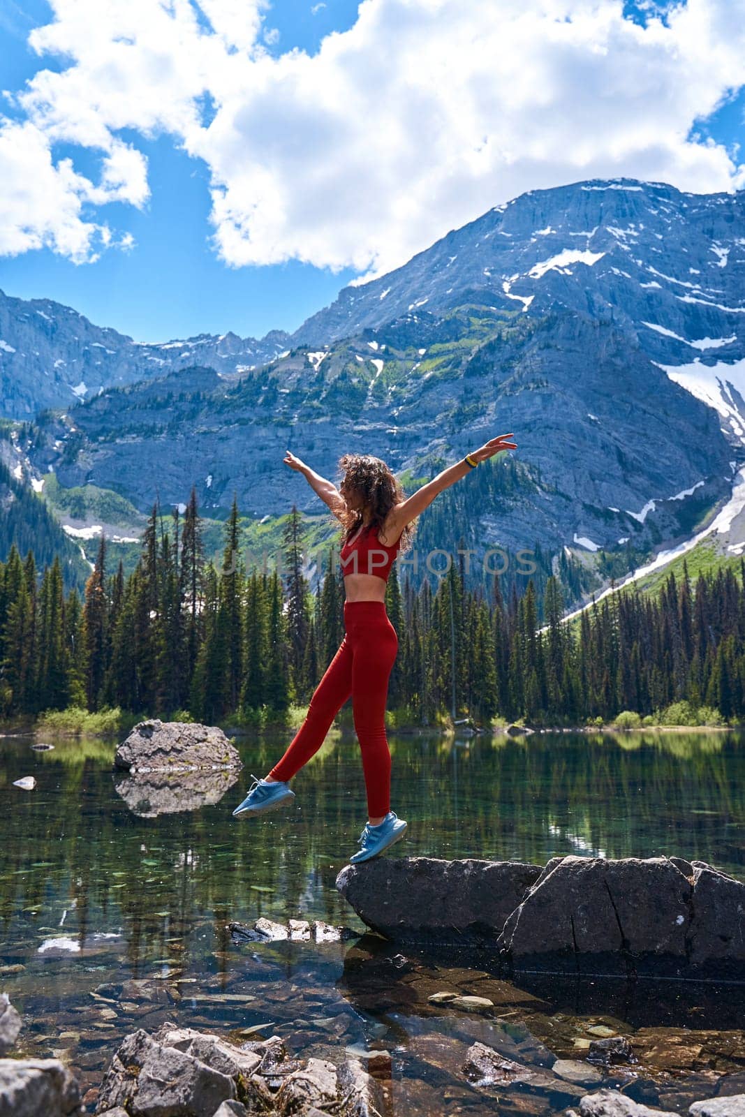 Beautiful slender woman in sports suit poses on big stone in calm clear lake with reflection of high mountains with snow in summer. Young girl resting by the water while hiking in Kananaskis Alberta. by Try_my_best