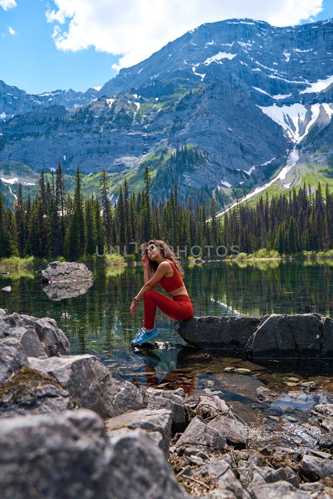 Beautiful slender woman in sports suit poses on big stone in calm clear lake with reflection of high mountains with snow in summer. Young girl resting by the water while hiking in Kananaskis Alberta