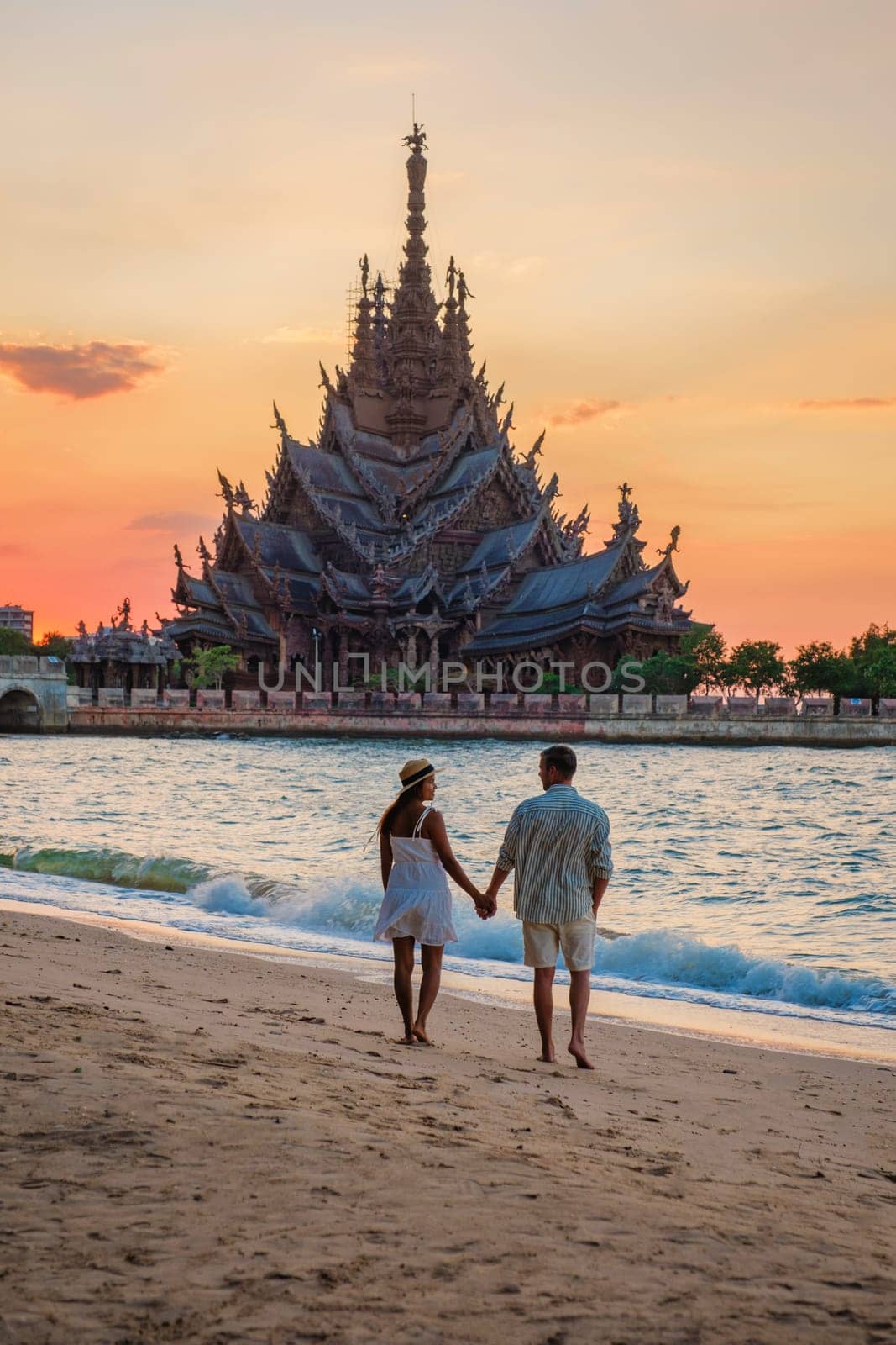 A diverse multiethnic couple of men and women visit The Sanctuary of Truth wooden temple in Pattaya Thailand at sunset on the beach