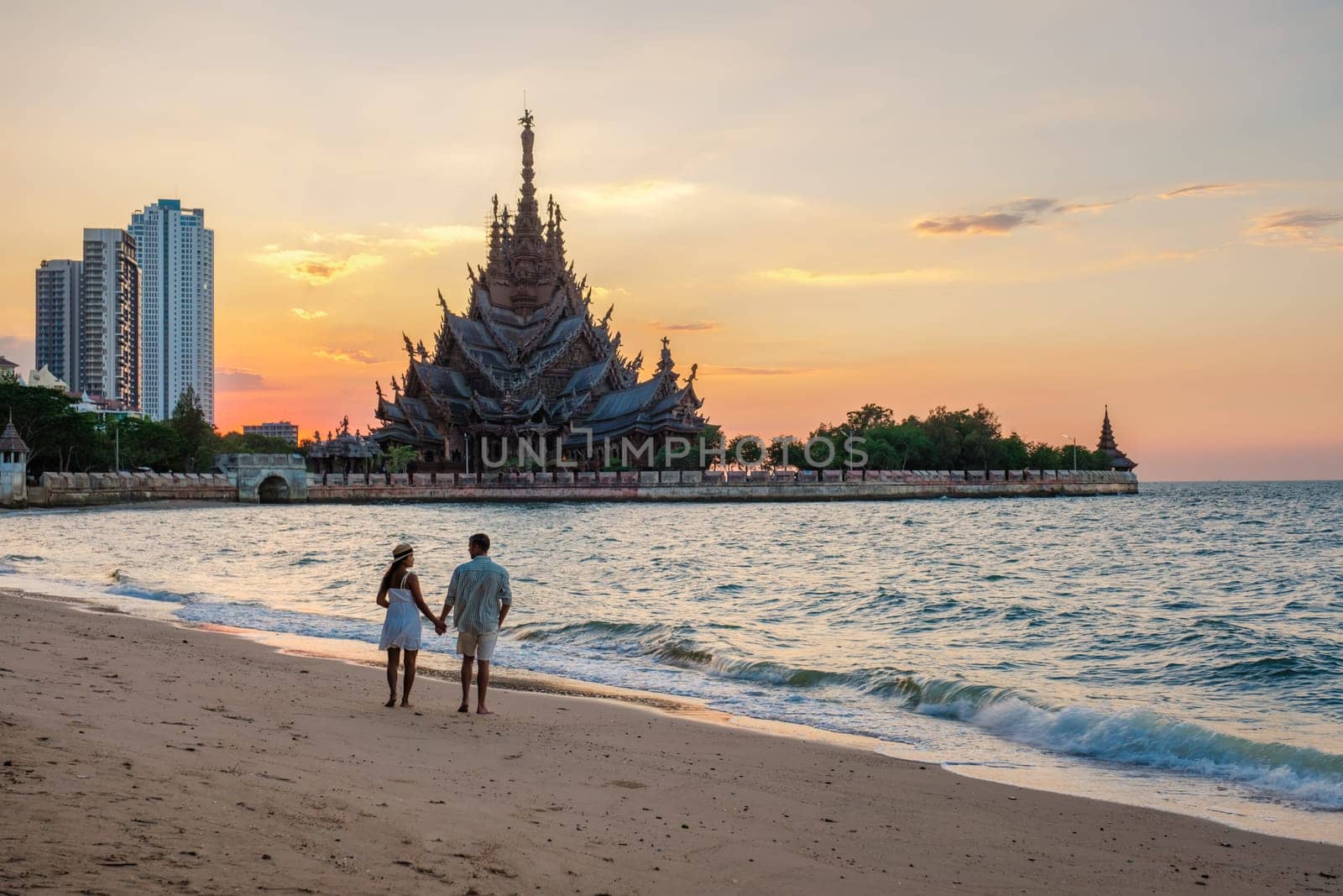 A diverse multiethnic couple of men and women visit The Sanctuary of Truth wooden temple in Pattaya Thailand at sunset on the beach