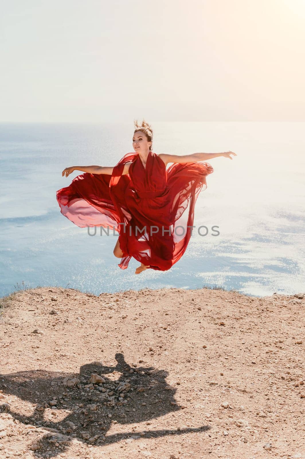 Side view a Young beautiful sensual woman in a red long dress posing on a rock high above the sea during sunrise. Girl on the nature on blue sky background. Fashion photo.