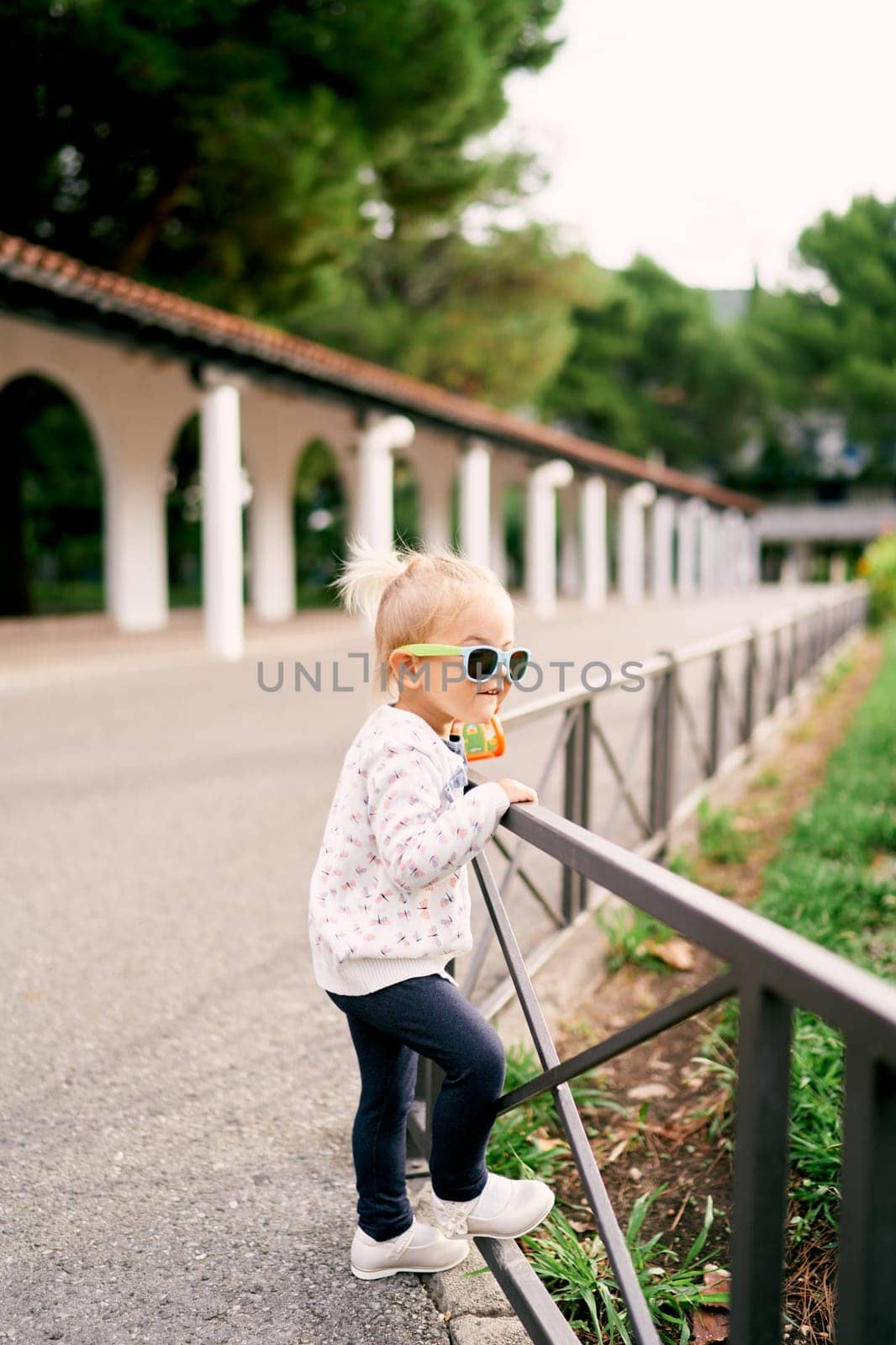 Little girl in sunglasses stands leaning on a fence and looks at the trees in the garden by Nadtochiy