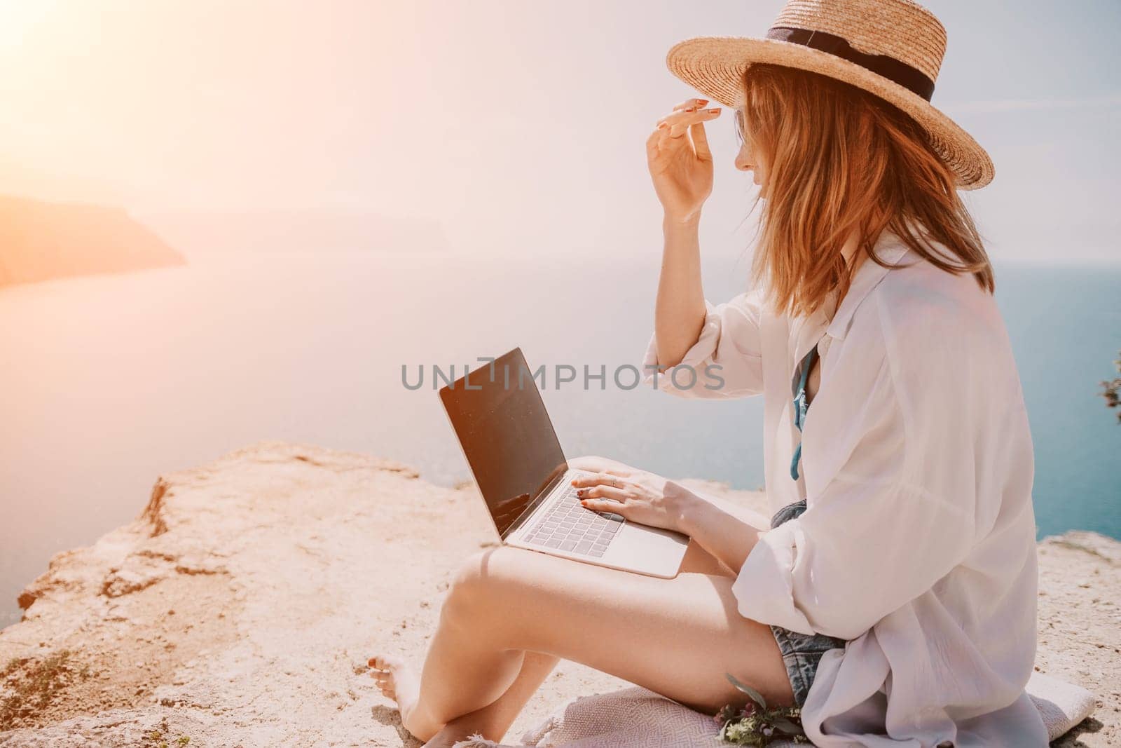 Successful business woman in yellow hat working on laptop by the sea. Pretty lady typing on computer at summer day outdoors. Freelance, travel and holidays concept.