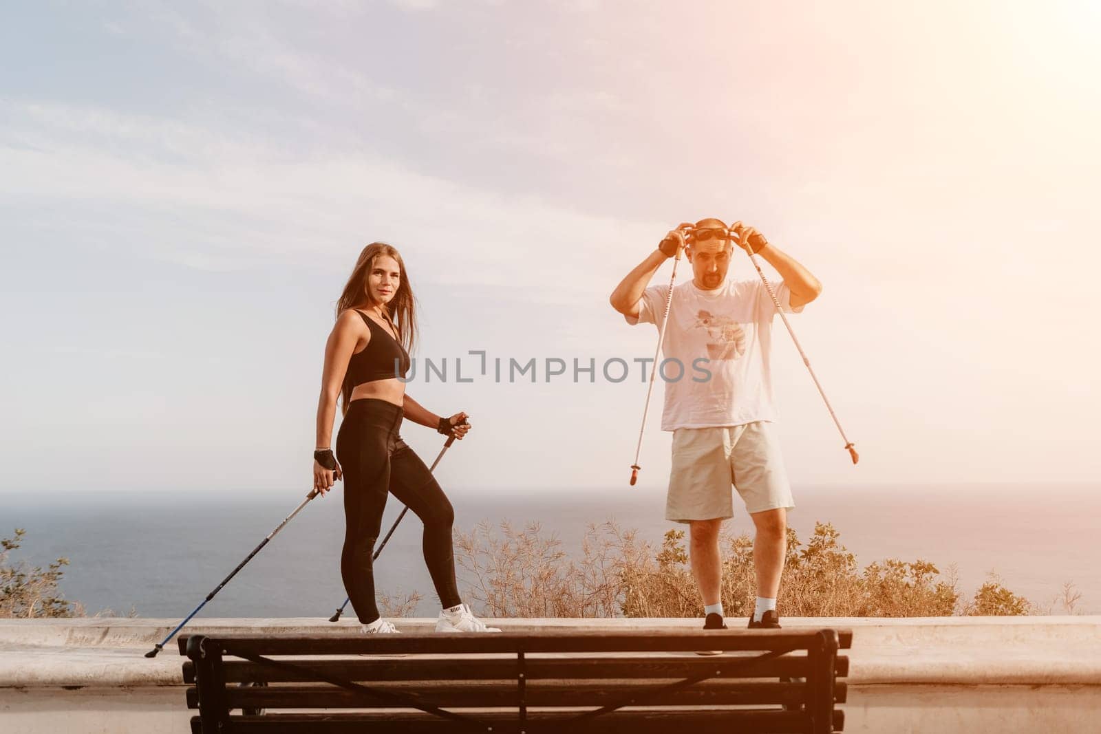 Happy Middle aged couple or friends practicing nordic walking in park near sea. Mature couple with trekking poles walking, practicing Nordic walking outdoors. Aging youthfully and sport concept by panophotograph