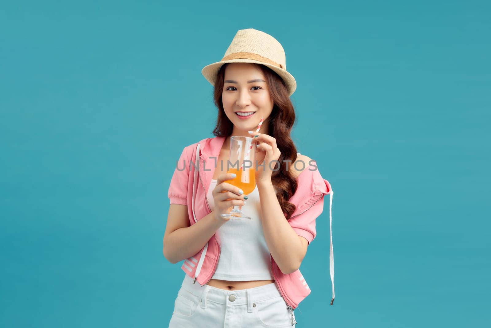Happy smiling young asian woman tourist drinking orange juice isolated on blue studio background by makidotvn