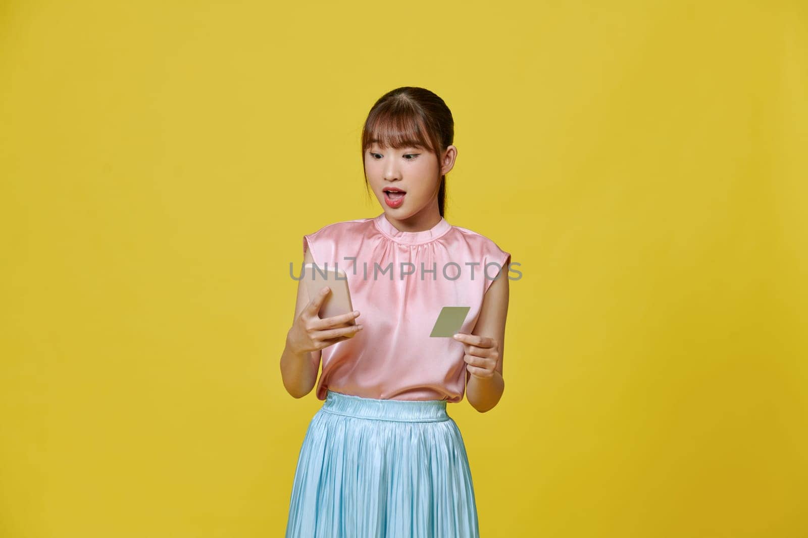 excited young lady isolated over yellow background using mobile phone holding credit card.