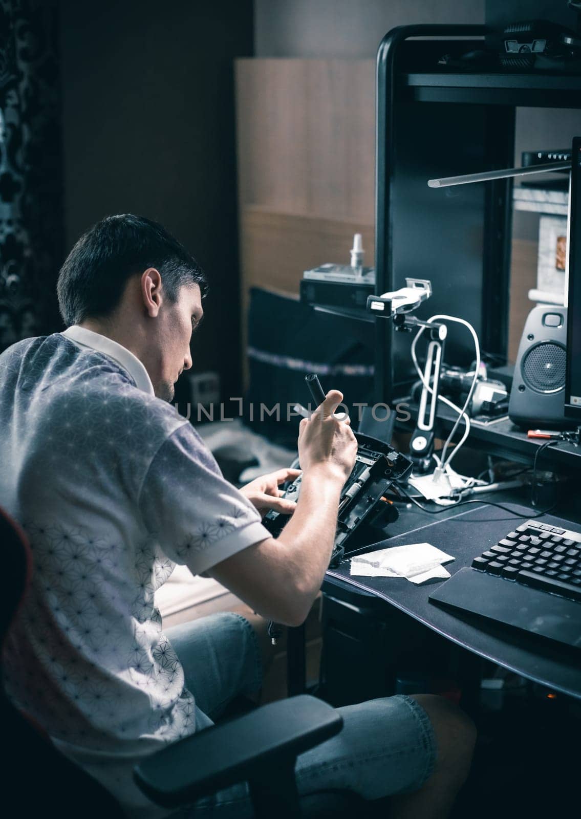 One young caucasian man repairing a motor control wheel on a joystick, connecting wires with a soldering iron, sitting in an armchair at a computer desk in a room, close-up side view.