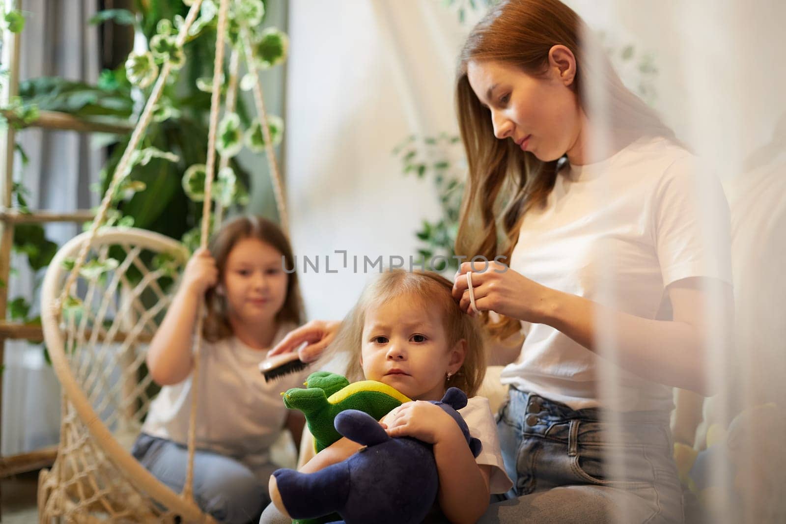 Young loving mom making ponytail to little preschool daughter, adult sister on background in living room. Mother helping child girl with hairstyle