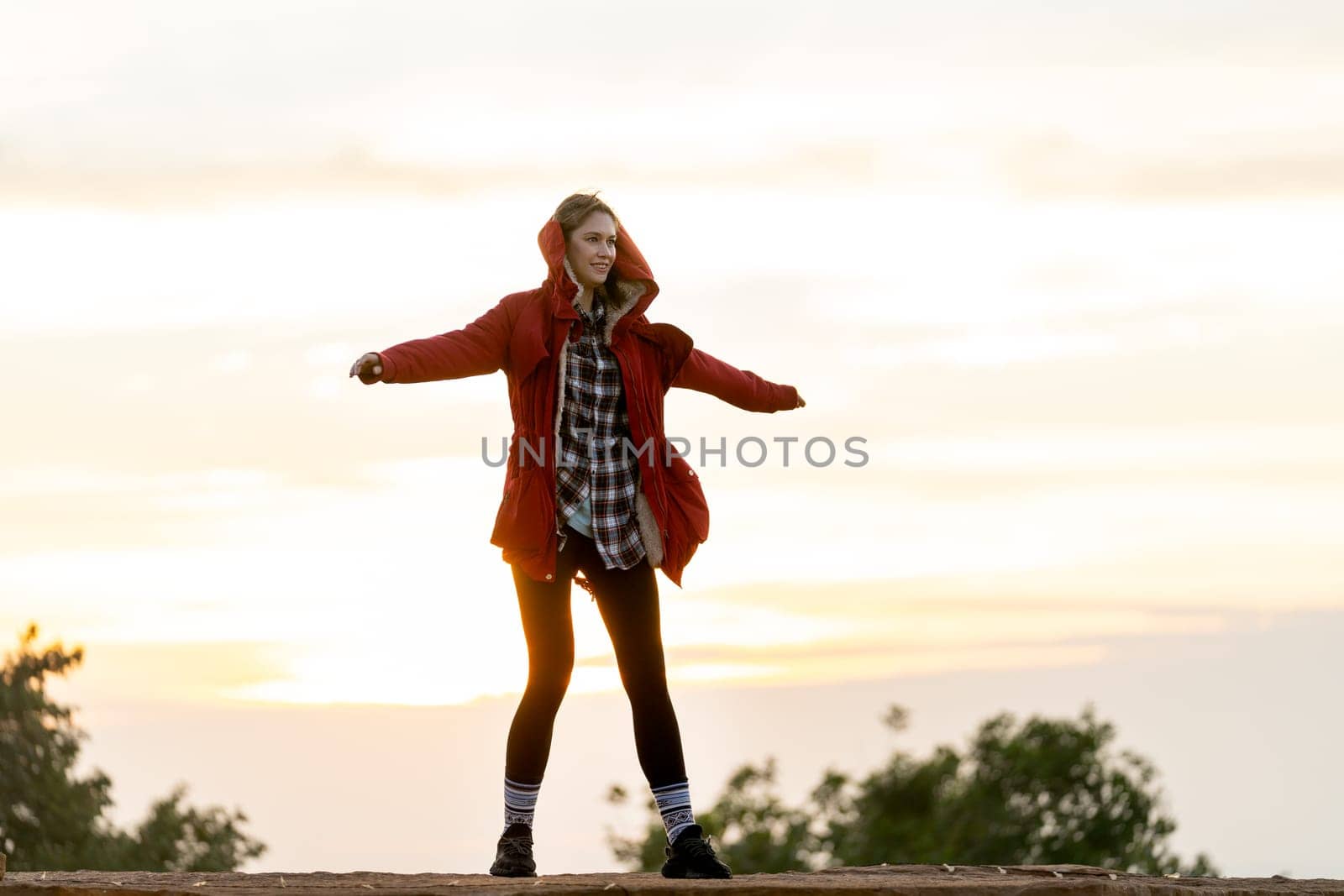 Beautiful Caucasian woman with red coat stand with spread her arms on the rock near the cliff on the mountain and background with warm light of sun rise in the morning.