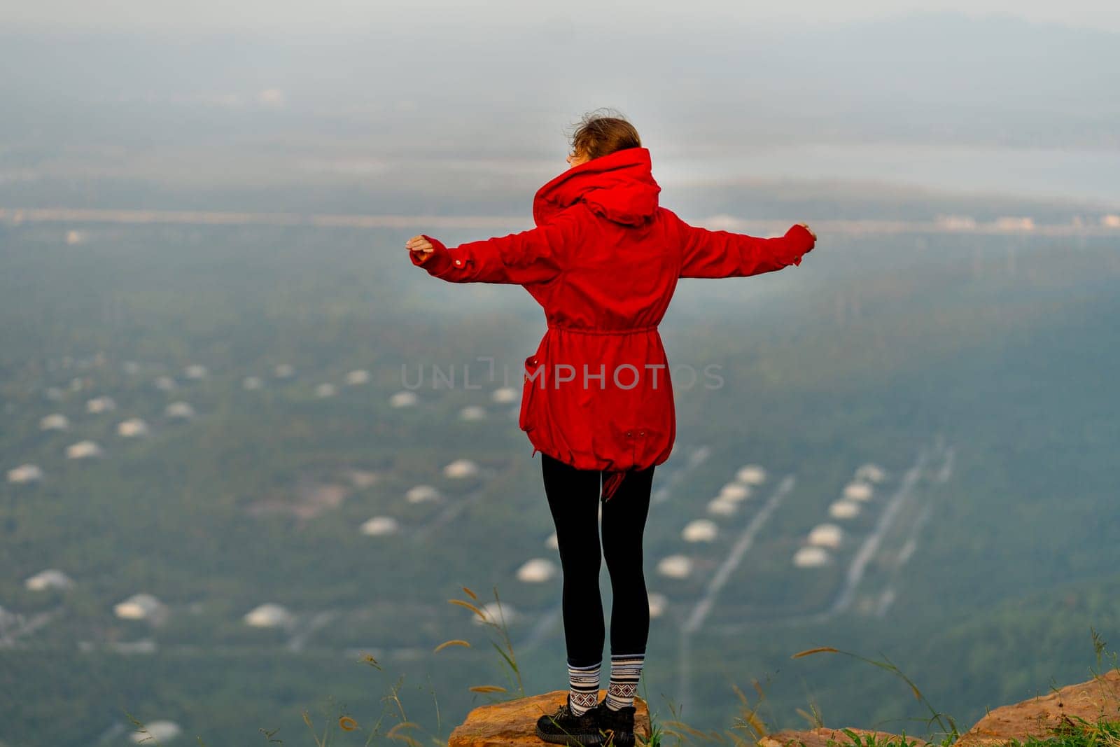 Back of woman wear red coat and stand with spread her arms near cliff on the mountain and look forward to beautiful view with warm light of sun rising.