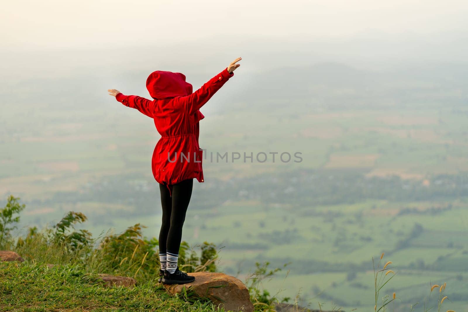 Back of woman wear red coat and cover with hood and stand with spread her arms near cliff on the mountain and look forward to beautiful view with warm light in the morning.