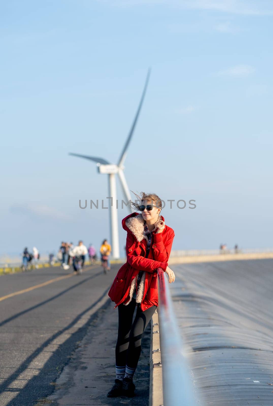 Vertical image of Caucasian waman with sunglasses stand in front of wind turbine or windmill also look to left side near the road.