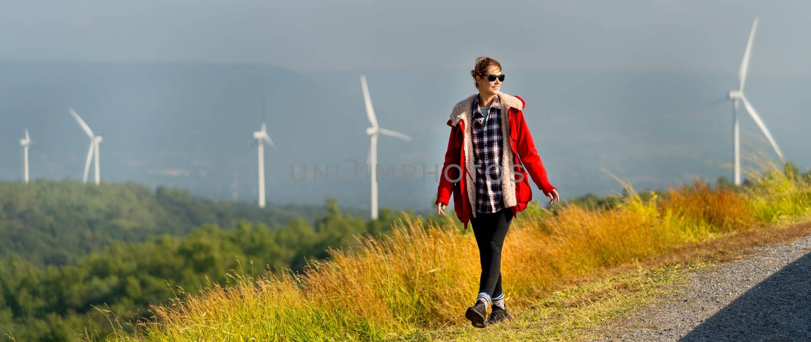 Caucasian woman with red coad and sunglasses walk along the roadside near the meadow and wind turbines or windmill with warm morning light. by nrradmin