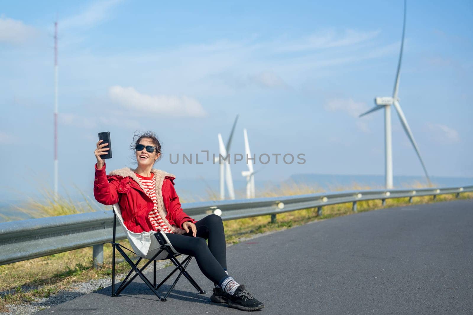 Caucasian woman with red coat and sunglasses sit on chair on roadside near wind turbines or windmill also use mobile phone to selfie with beautiful view on the mountain.