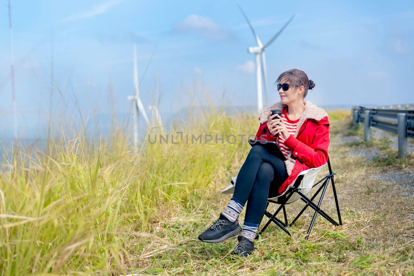Caucasian woman sit on chair near meadow with blue sky and wind turbines or windmill on the back and she also hold a cup of coffee with happiness.