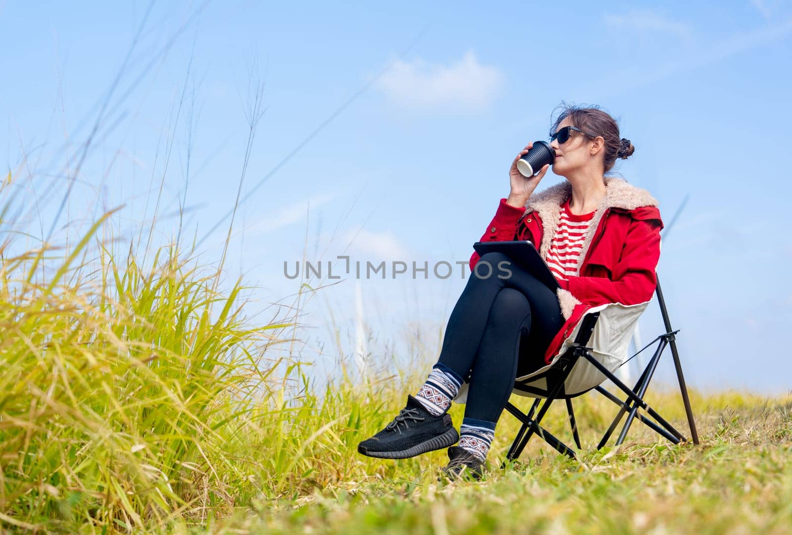 Caucasian woman sit on chair near meadow with blue sky and drink coffee with relax action in front of wind turbines or windmill.
