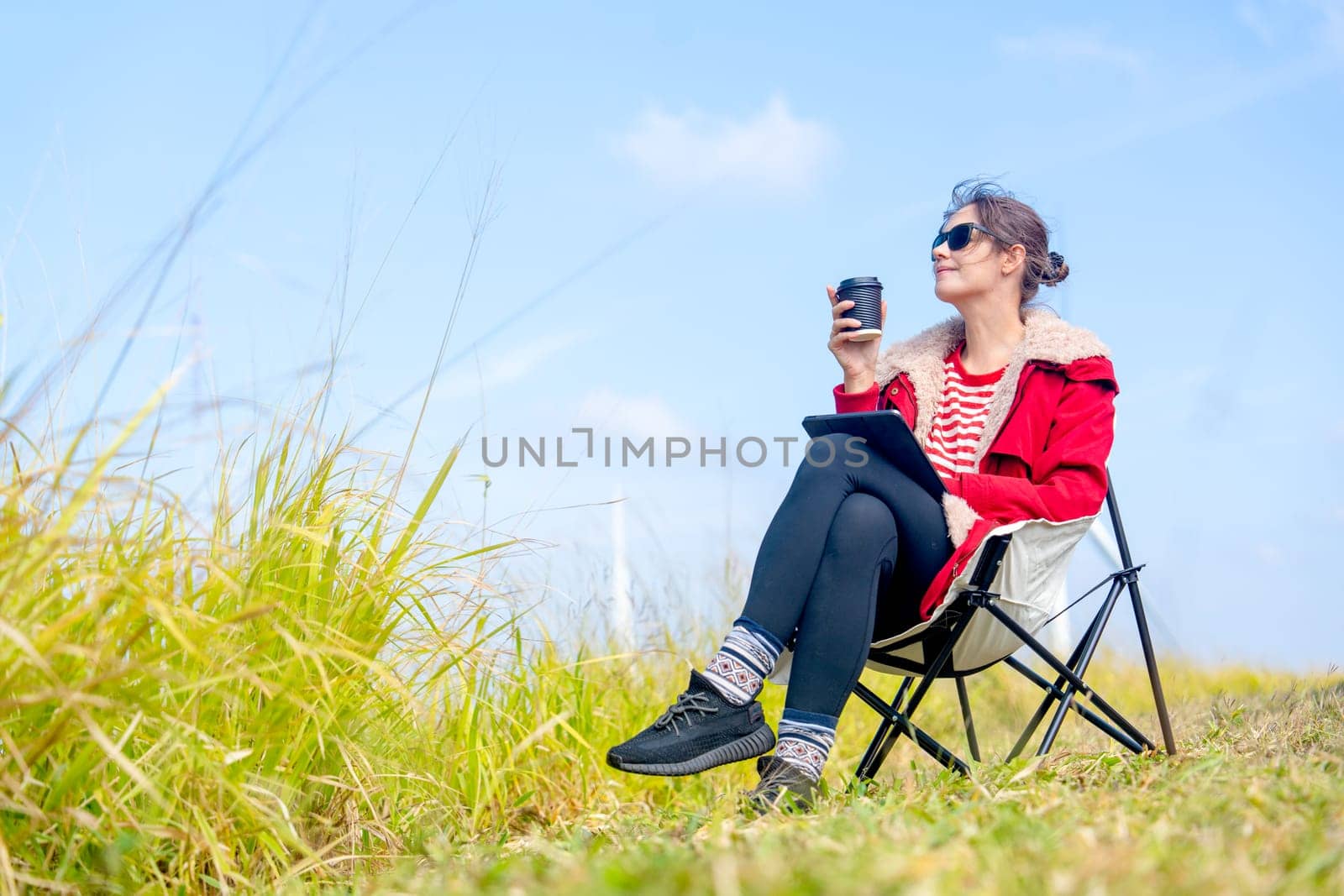 Beautiful Caucasian woman sit on chair near meadow with blue sky and wind turbines or windmill on the back and she also hold a cup of coffee with relax action.