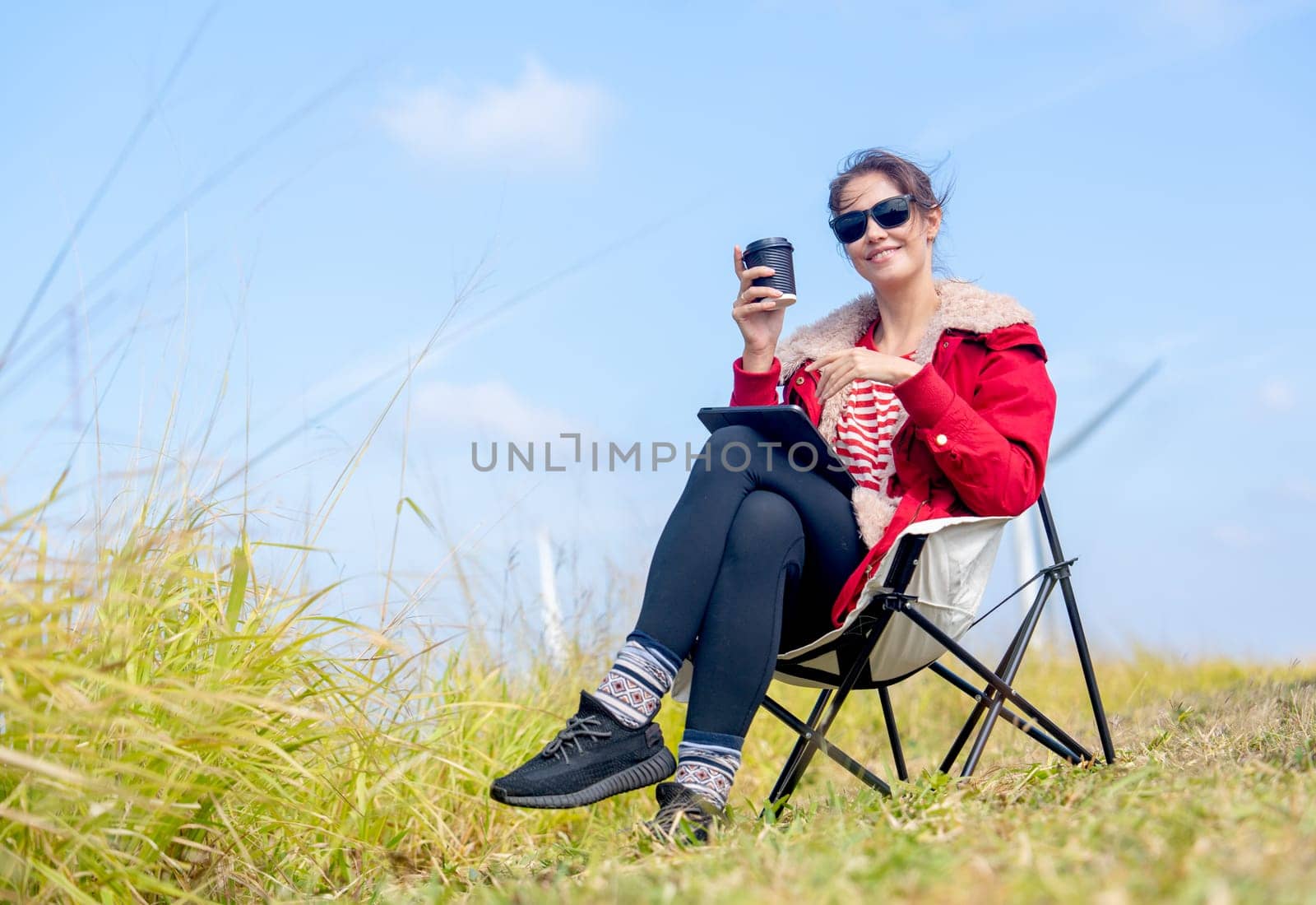 Caucasian woman sit on chair near meadow with blue sky and wind turbines or windmill on the back and she also hold a cup of coffee and look at camera.