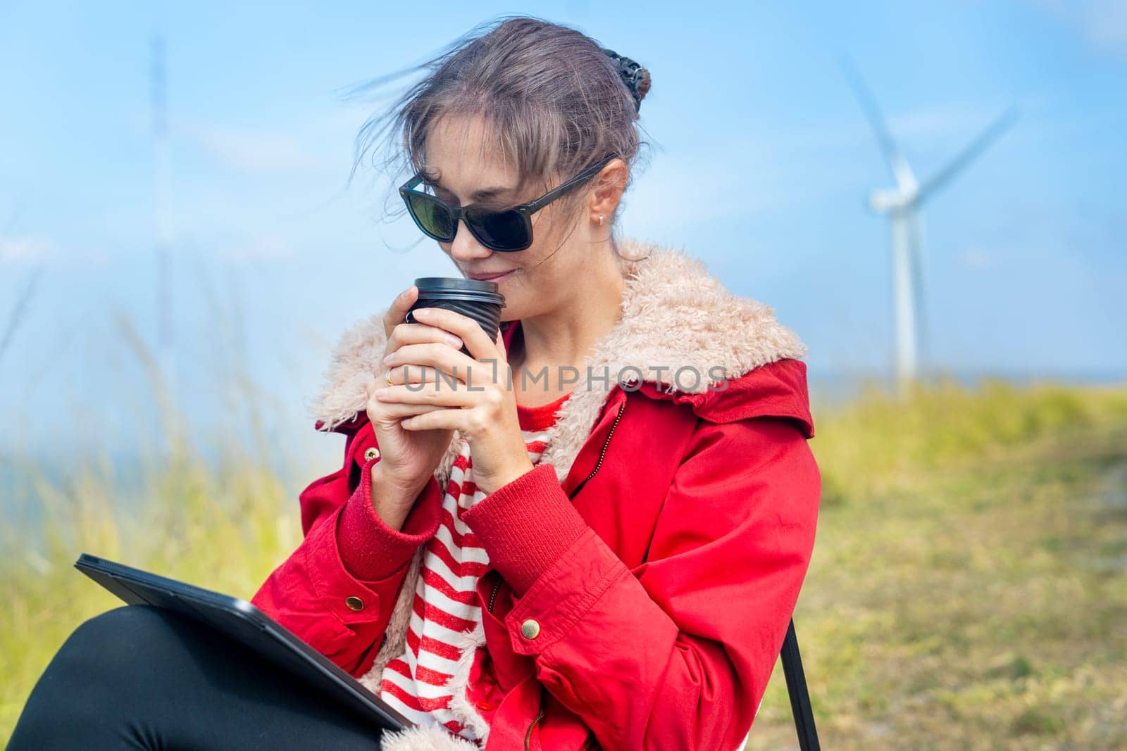 Close up Caucasian woman hold a cup of coffee and drink near meadow and wind turbines on the background.