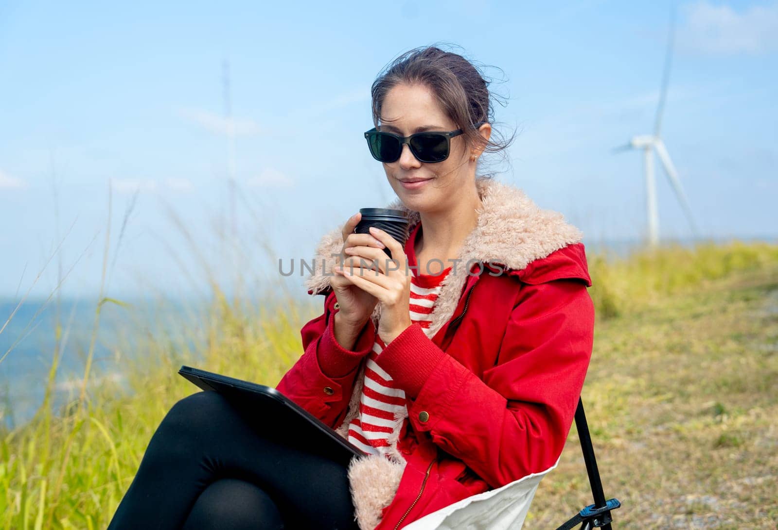 Close up Caucasian woman hold a cup of coffee and look at the camera and sit near meadow and wind turbines on the background.