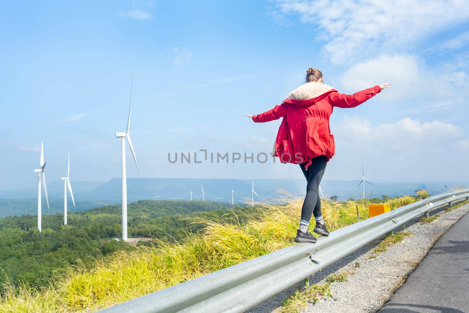 Back of woman with red coat walk on barrier fence of sideroad near wind turbines or windmill on the mountain.