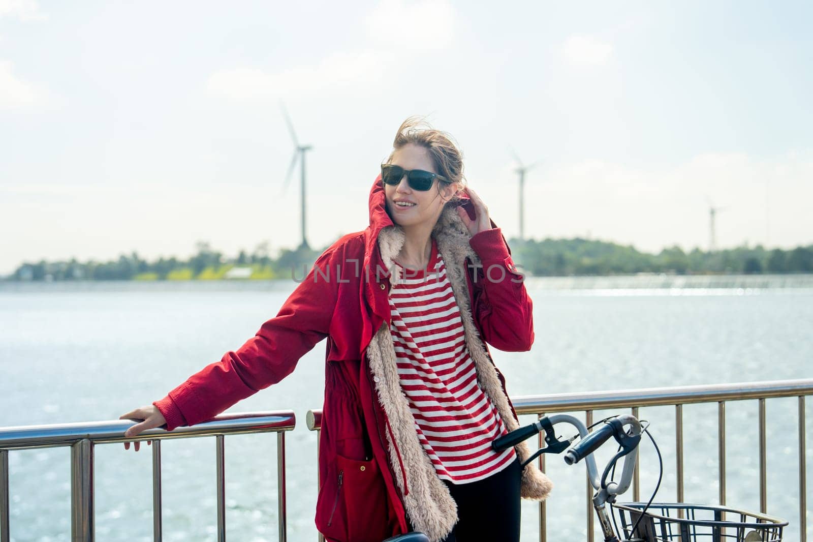 Close up Woman with red coat and sun glasses lean to fence and touch coat hood also hold hand of bicycle stand in front of wind turbines or windmill near water reservoir.