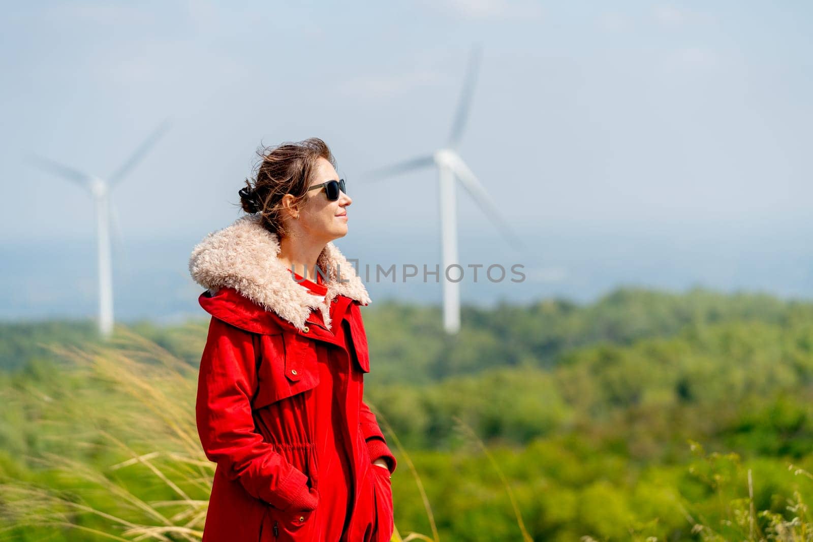 Caucasian woman with red coat and sunglasses stand with smiling and look to right side in front of wind turbines or windmill on the mountain.