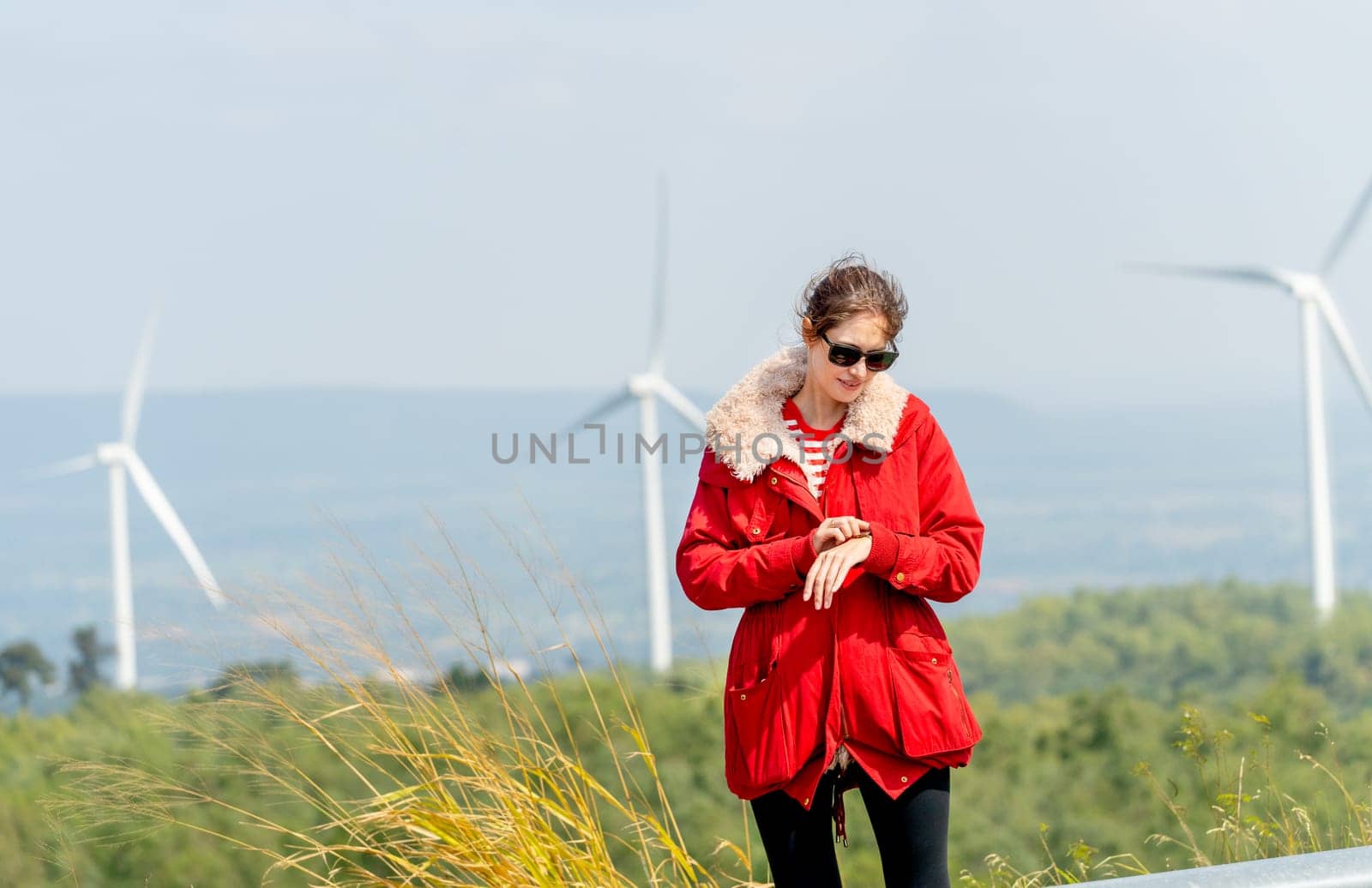 Caucasian woman with red coat look at her watch and stay on mountain with windmill or wind turbine as background.