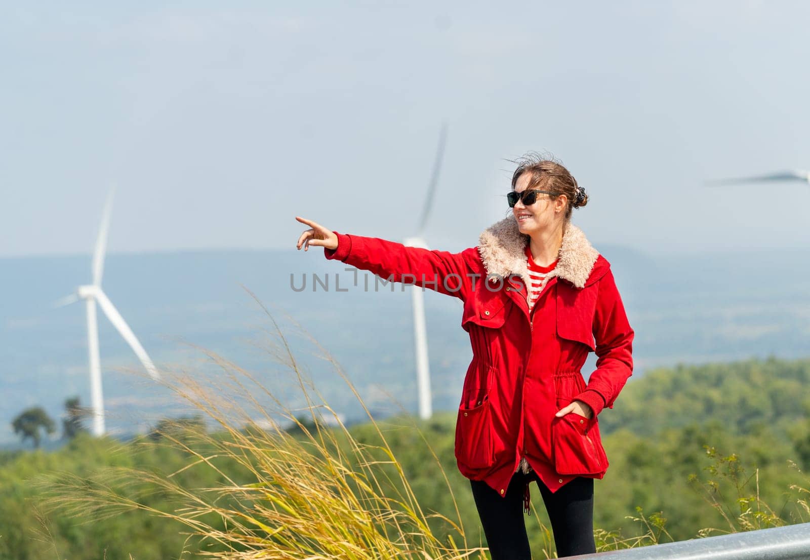 Caucasian woman with red coat stand with smiling and point to left side in front of wind turbines or windmill on the mountain.