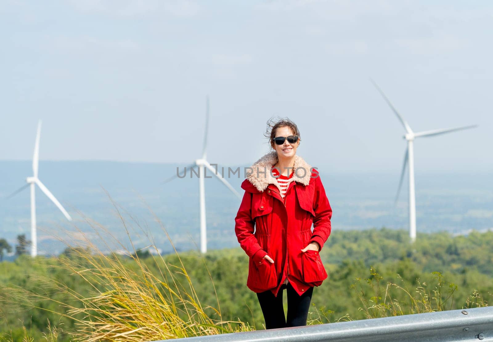Front view of Caucasian woman with red coat stand with look at camera on mountain near windmill or wind turbine in the back.