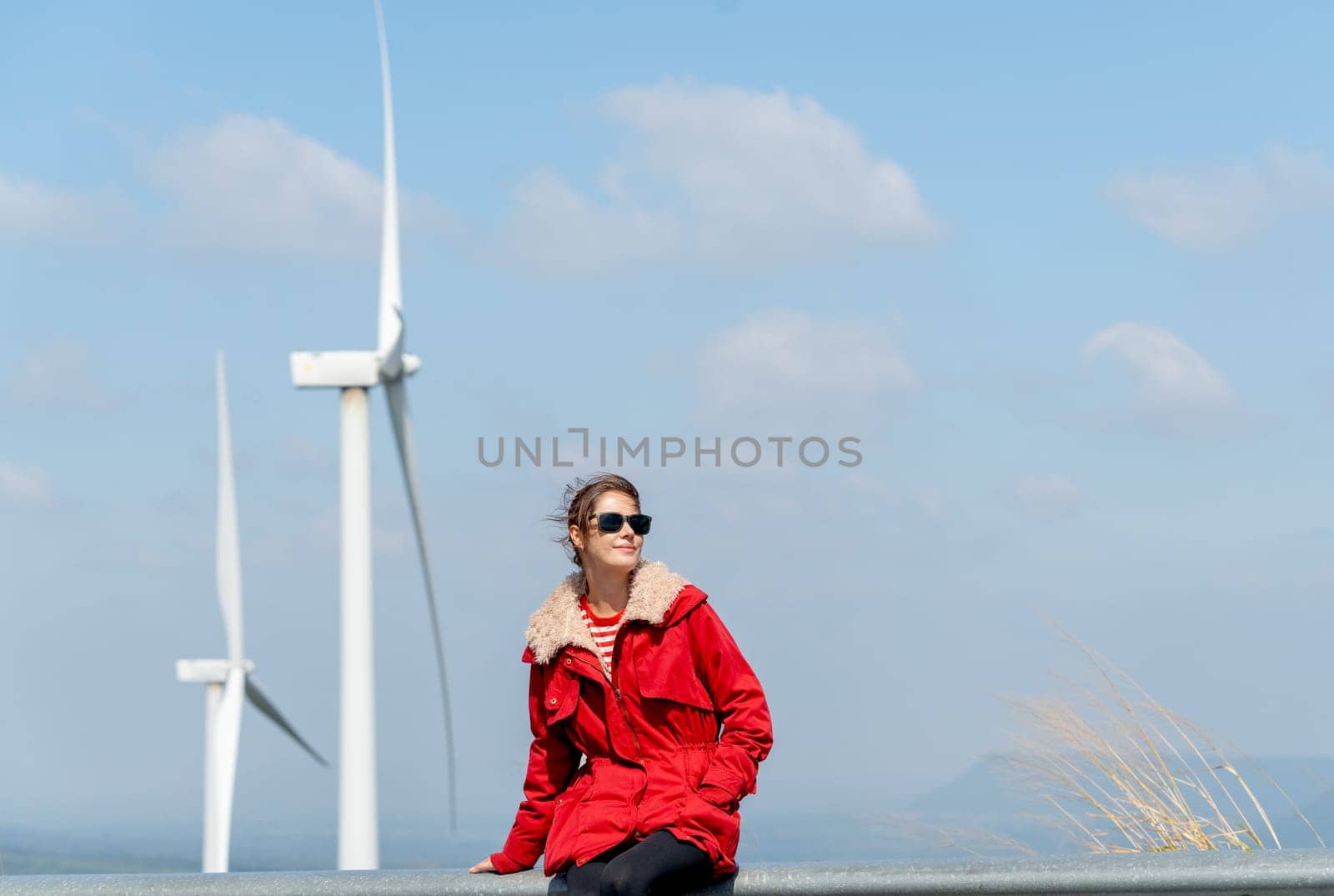 Front view of Caucasian woman with red coat sit on barrier of roadside and use mobile phone on mountain near windmill or wind turbine also look to right side.