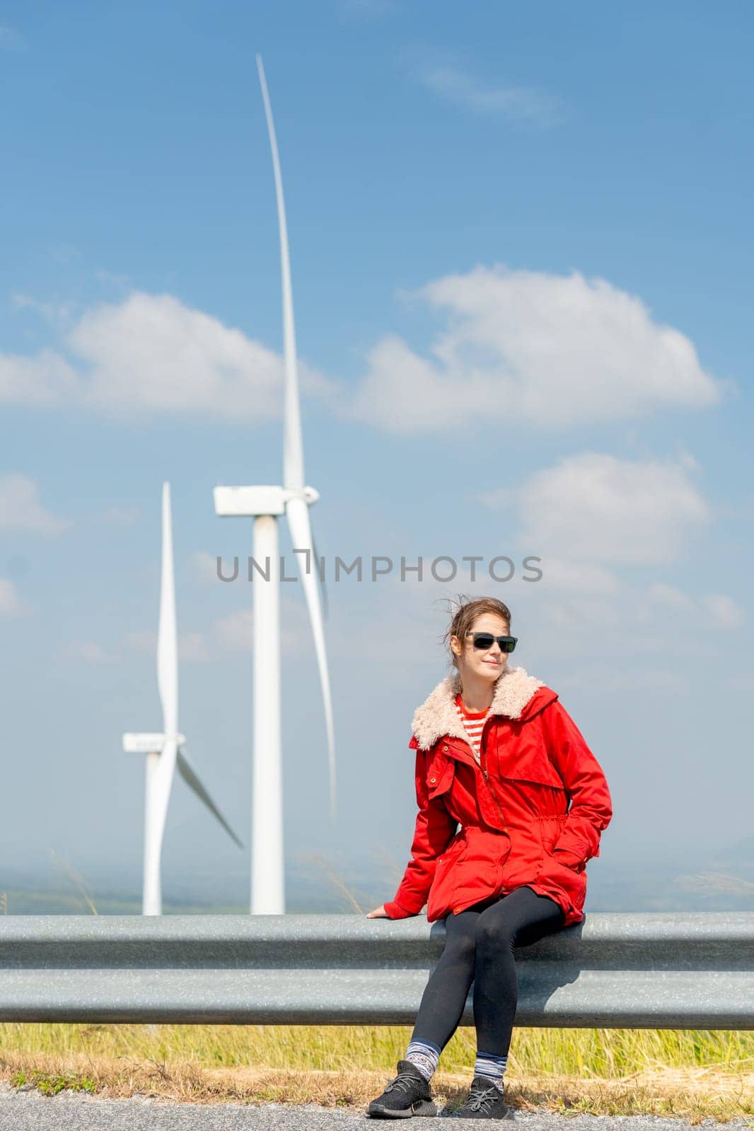 Vertical image of Caucasian woman with red coat sit on barrier of roadside and use mobile phone on mountain near windmill or wind turbine also look to right side.