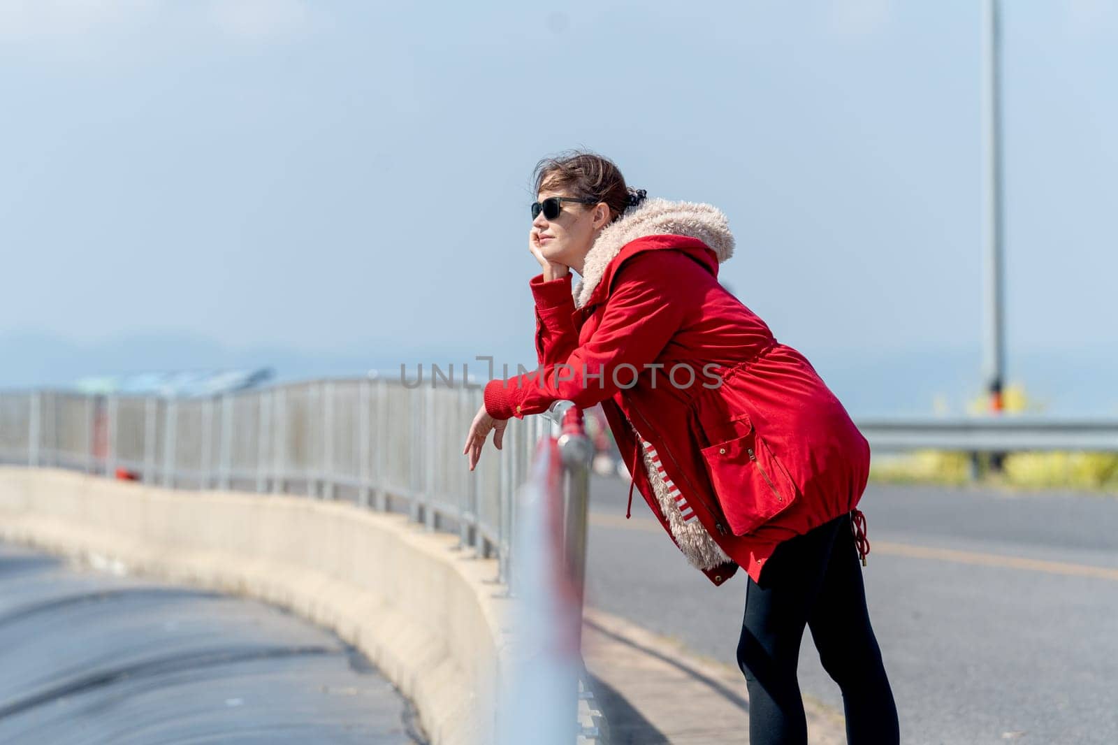 Side view of Caucasian woman with red coat and sun glasses lean to fence and look forward with day light and look cool.