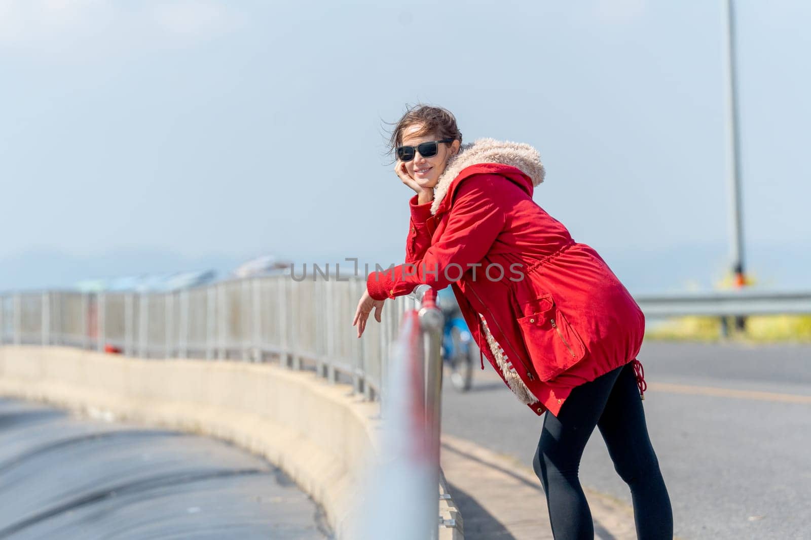 Woman with red coat and sun glasses lean to fence and look at camera with smiling with day light and look cool.