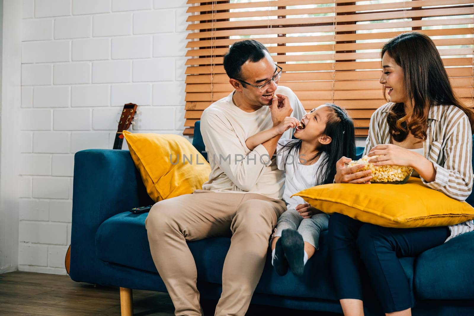 A happy family filled with joy watches TV while eating popcorn on modern sofa at home by Sorapop