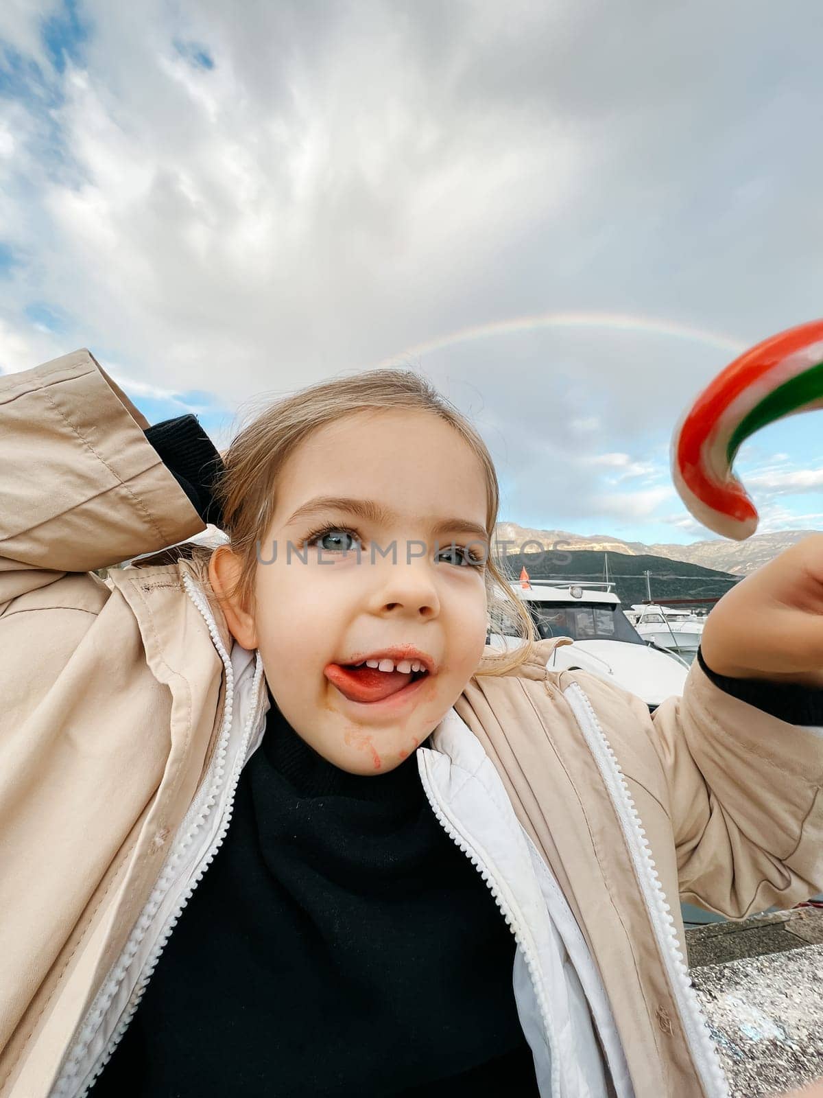 Little girl with her tongue hanging out and a candy in her hand against the backdrop of a rainbow. High quality photo