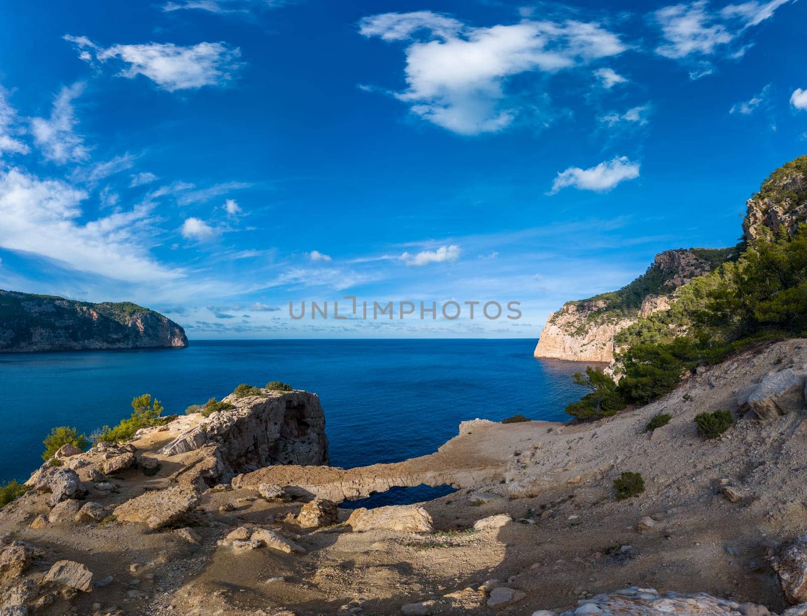 Cliffs rise above serene blue sea beneath a partly cloudy sky.