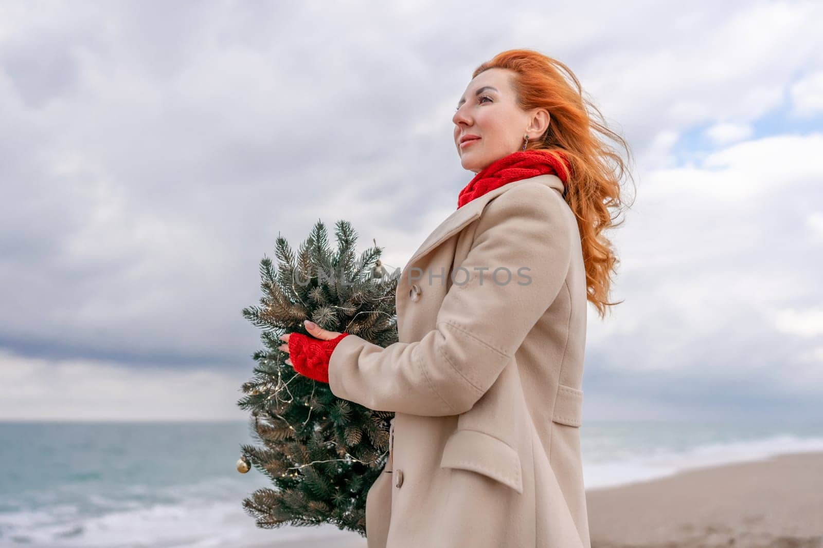 Redhead woman Christmas tree sea. Christmas portrait of a happy redhead woman walking along the beach and holding a Christmas tree in her hands. Dressed in a light coat, white suit and red mittens. by Matiunina