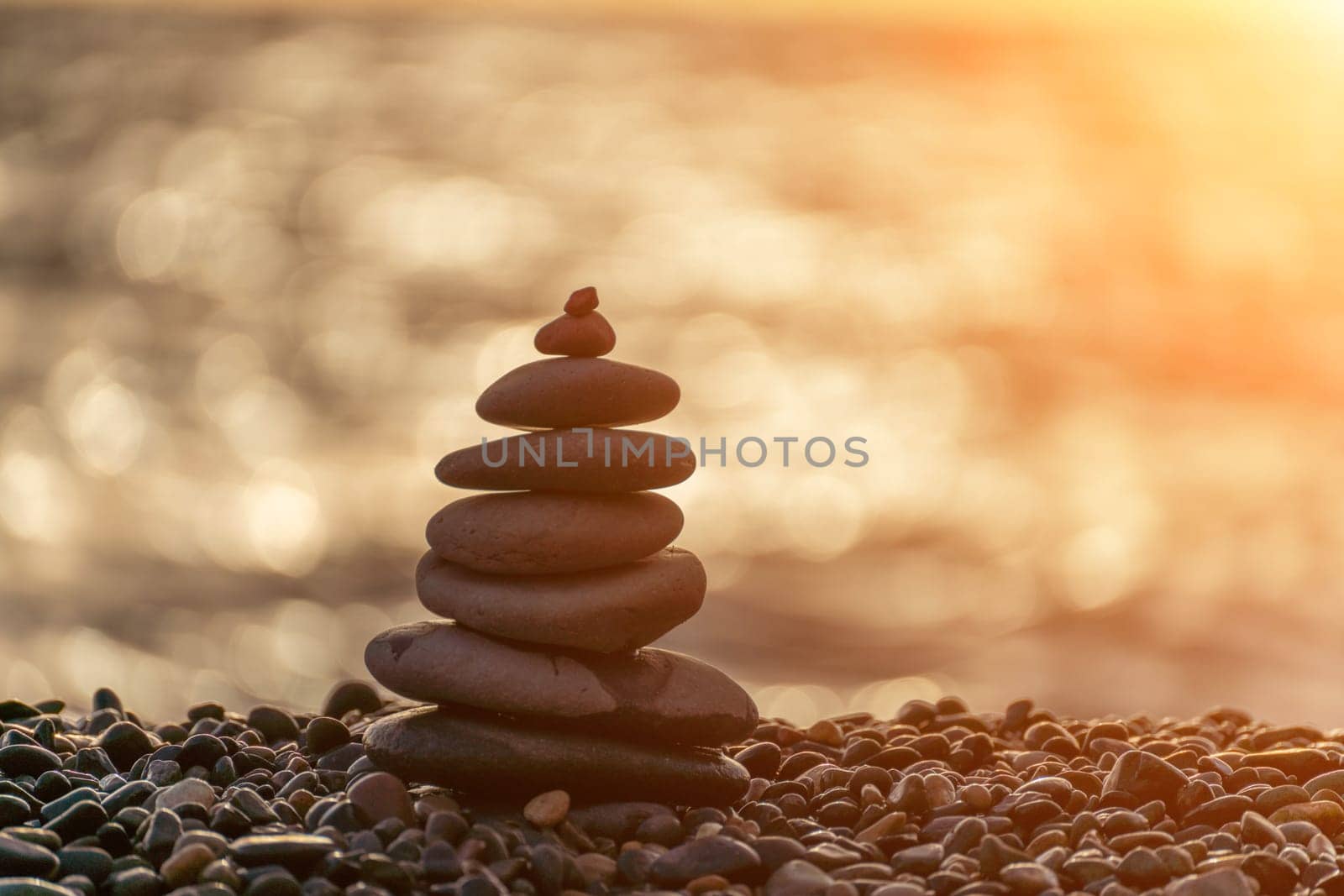 Balanced rock pyramid on pebbles beach. Golden sea bokeh on background. Selective focus, zen stones on sea beach, meditation, spa, harmony, calm, balance concept. by Matiunina