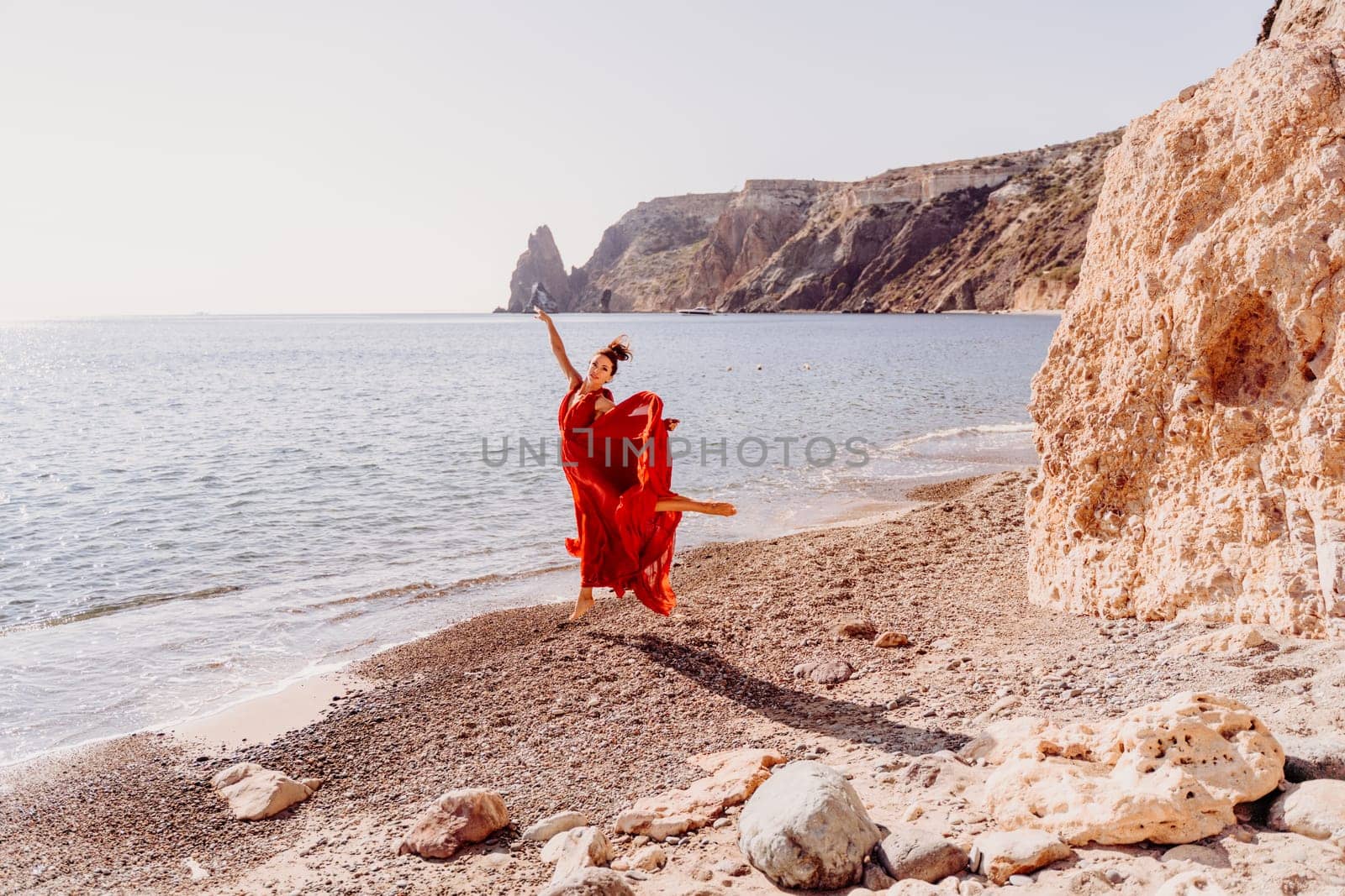 Woman red dress sea. Female dancer in a long red dress posing on a beach with rocks on sunny day. Girl on the nature on blue sky background