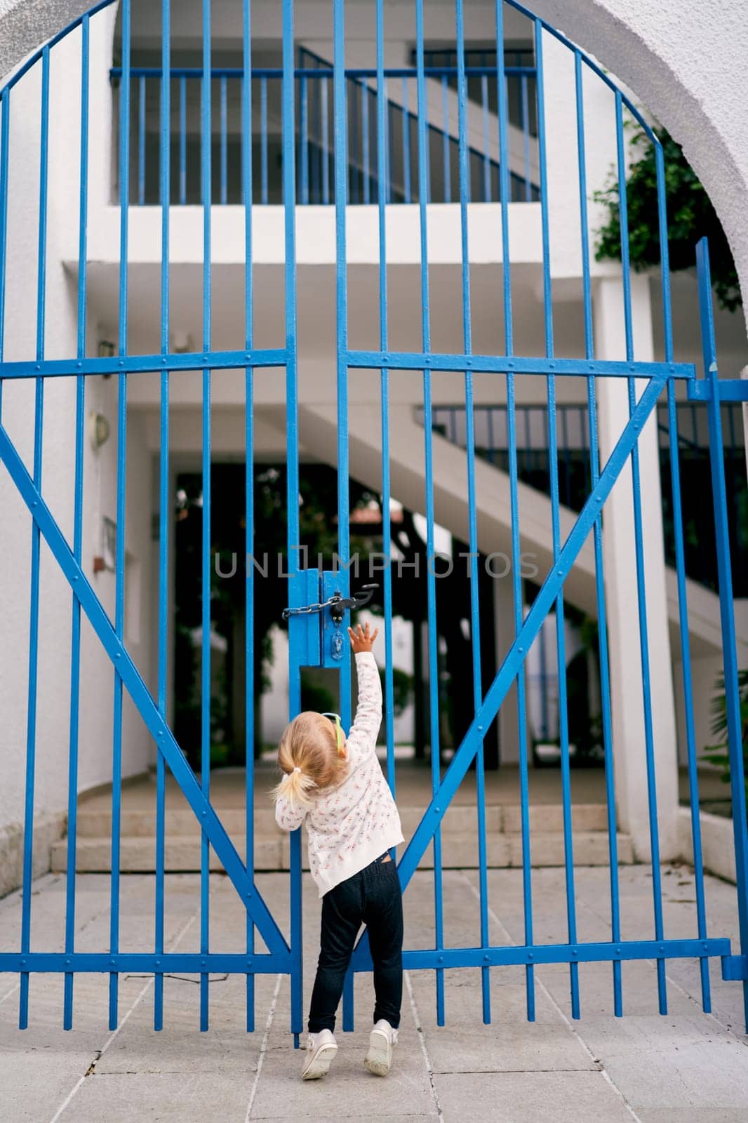 Little girl reaches for the lock on the metal arched gate of the house. Back view. High quality photo