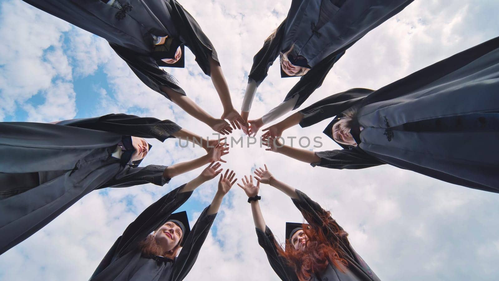 Team of college or university students celebrating graduation. Group of happy successful graduates in academic hats and robes standing in circle and putting their hands together.