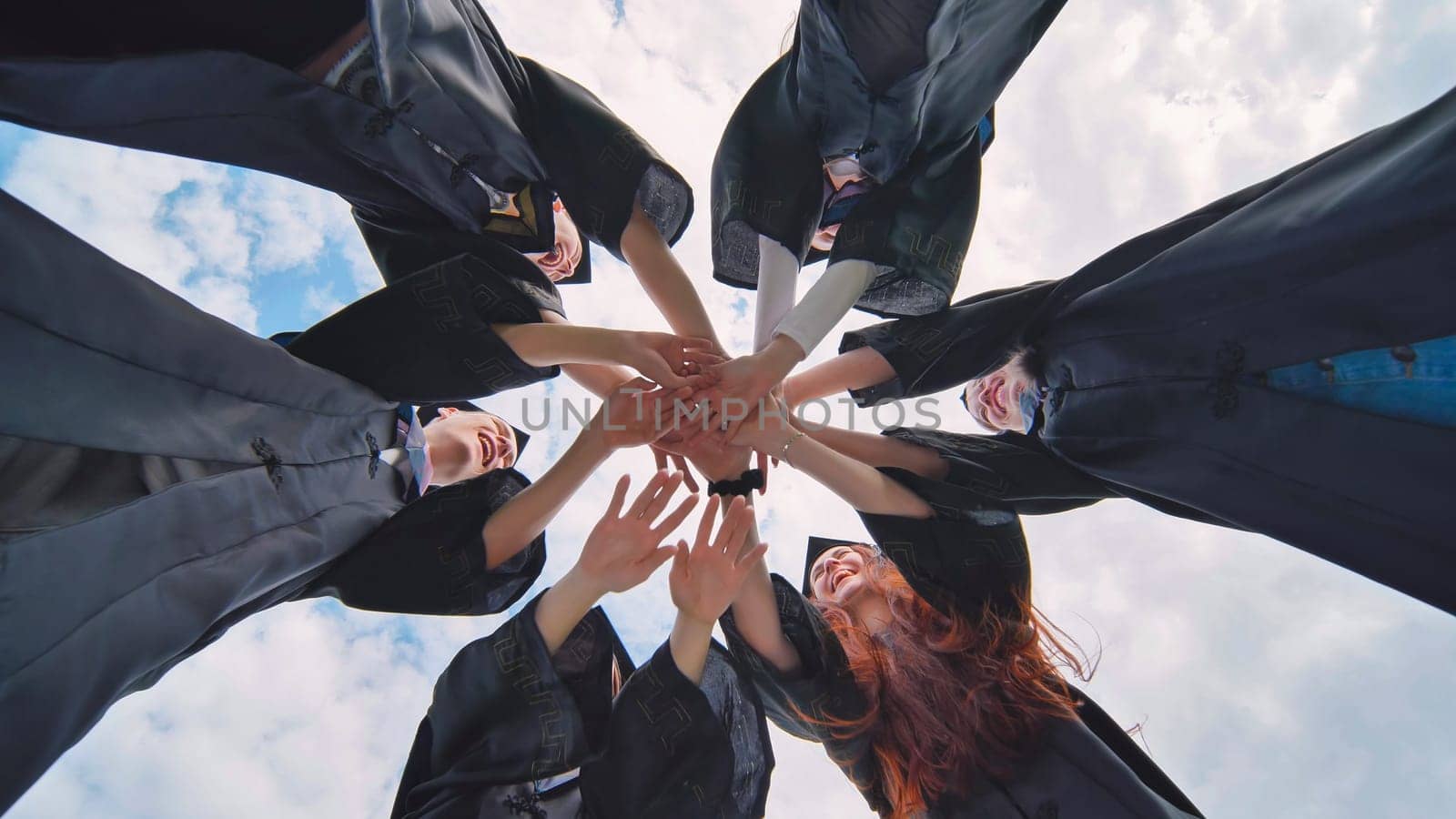 Team of college or university students celebrating graduation. Group of happy successful graduates in academic hats and robes standing in circle and putting their hands together