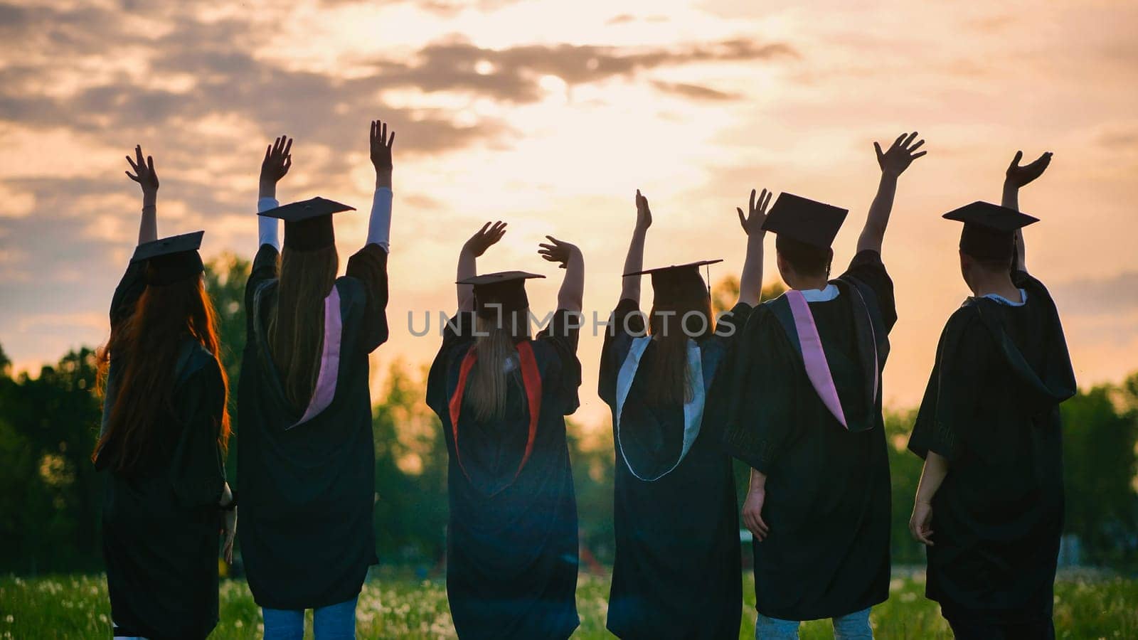 Silhouettes of graduates in black robes waving their arms against the evening sunset