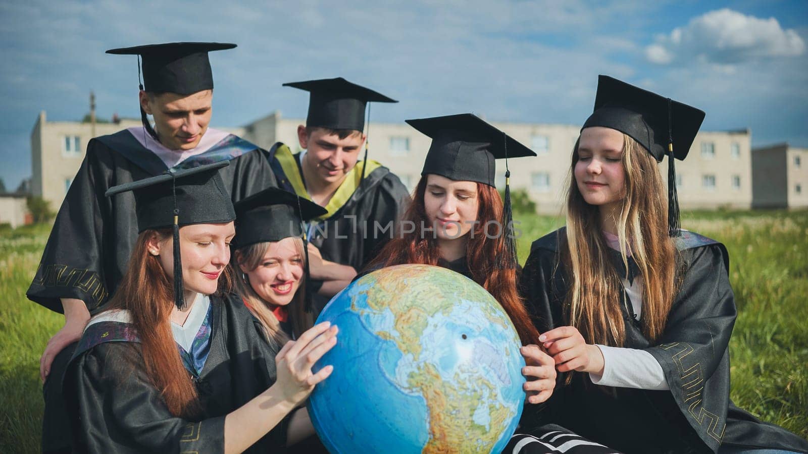 Graduates in black robes examine a geographical globe sitting on the grass. by DovidPro