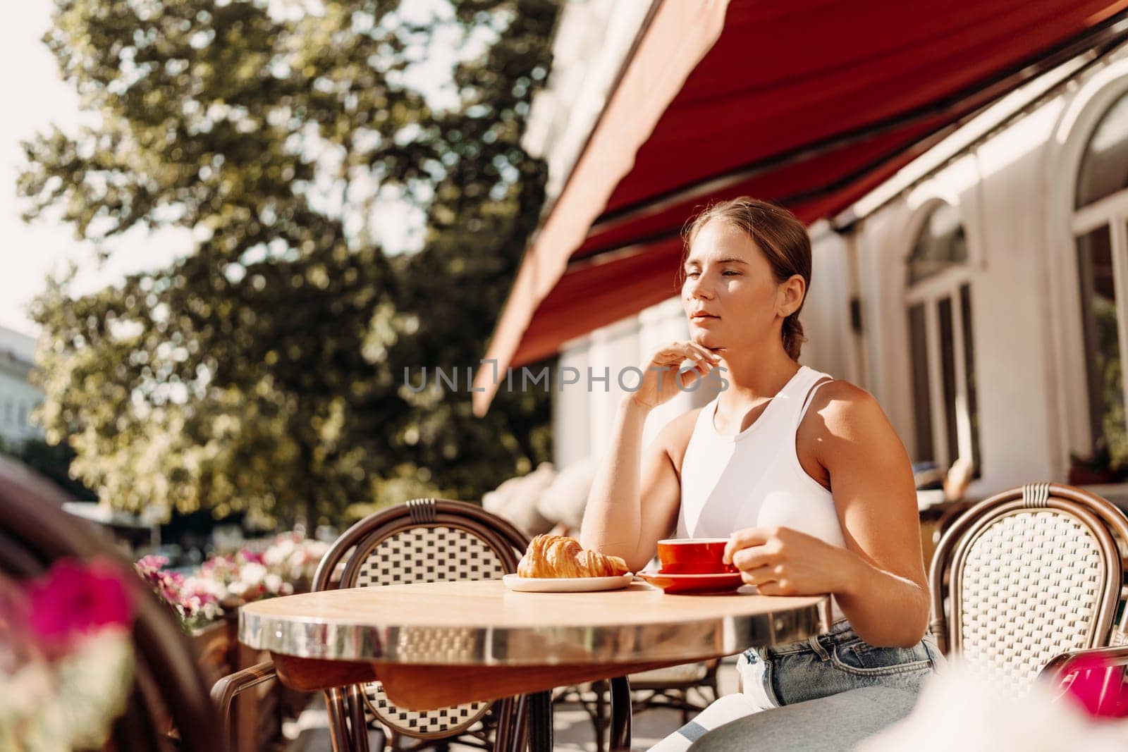 Portrait of happy woman sitting in a cafe outdoor drinking coffee. Woman while relaxing in cafe at table on street, dressed in a white T-shirt and jeans by Matiunina